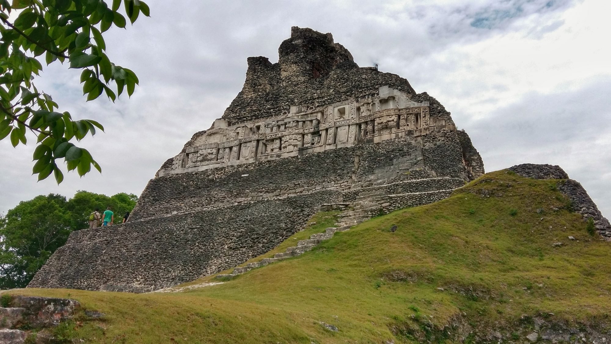Ancient Mayan pyramid with carved stone decorations and stairs, surrounded by green grass and trees, under a cloudy sky.