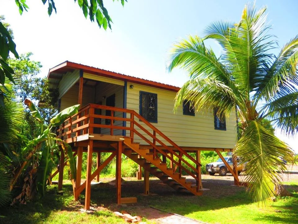 A small house on stilts painted yellow with red trim, surrounded by lush tropical plants and palm trees, with a wooden staircase leading to a front porch, under a clear blue sky.