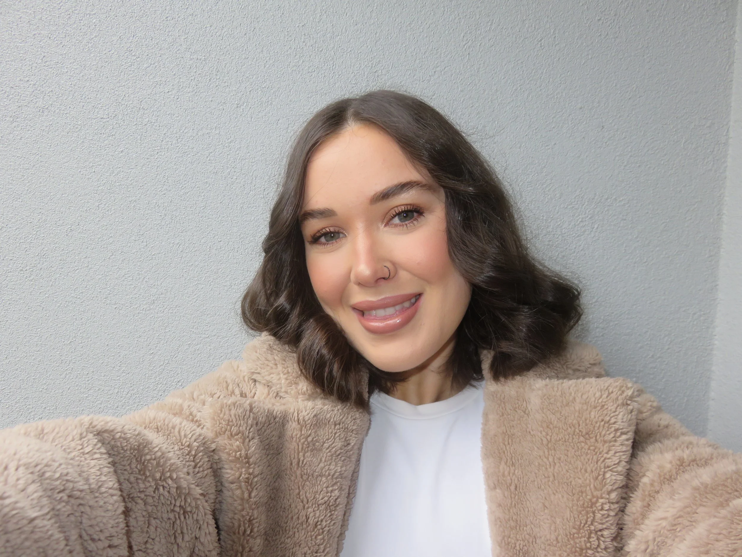 A young woman with curly brown hair, blue eyes, and a nose ring smiling and taking a selfie against a plain gray wall. She is wearing a beige fuzzy jacket over a white shirt.