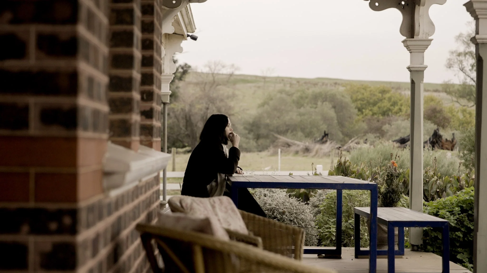 A woman with black hair and glasses sitting at a table on a porch, looking out at a rural landscape with trees and open fields in the distance.