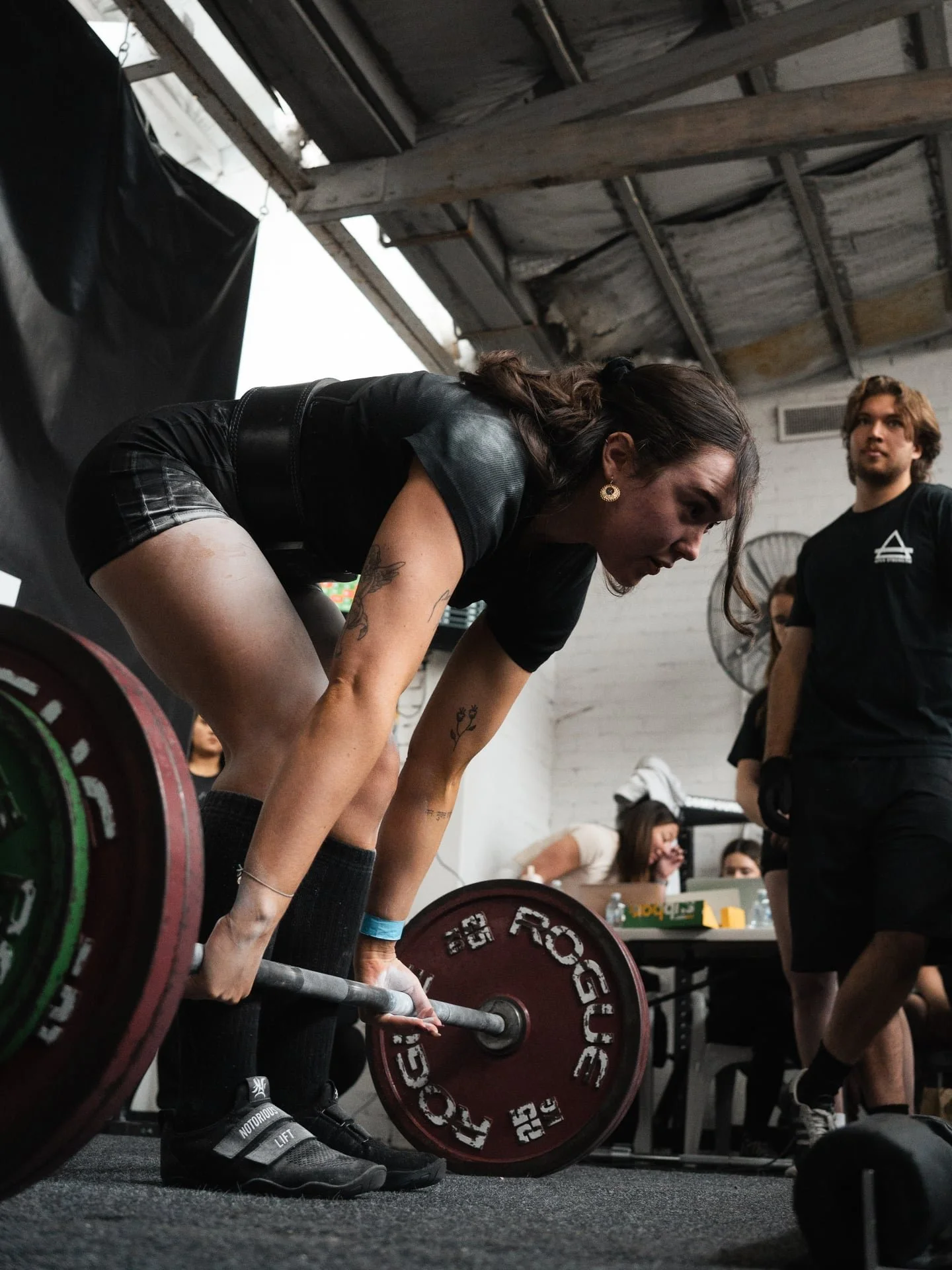 A woman with tattoos and dark hair prepares to lift a barbell loaded with red and green weight plates during a weightlifting competition, with a focused expression on her face. Other people are seen in the background, including a person in black attire and spectators seated at a table.