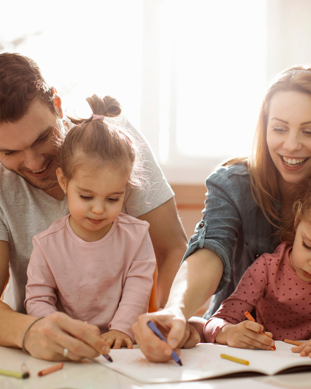 A happy family with two adults and two young girls drawing together at a table.