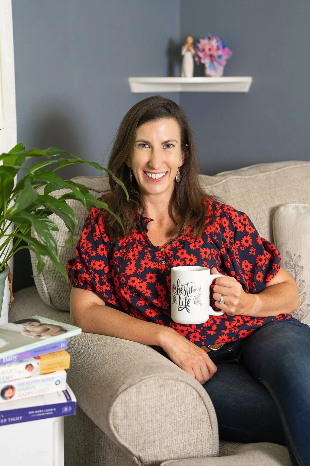 A woman with shoulder-length brown hair, wearing a red and navy floral blouse, smiling while sitting on a beige couch with a white mug that says 'the best things in life are not things' in her hand, in a room with a gray wall, a potted plant, and a stack of books.