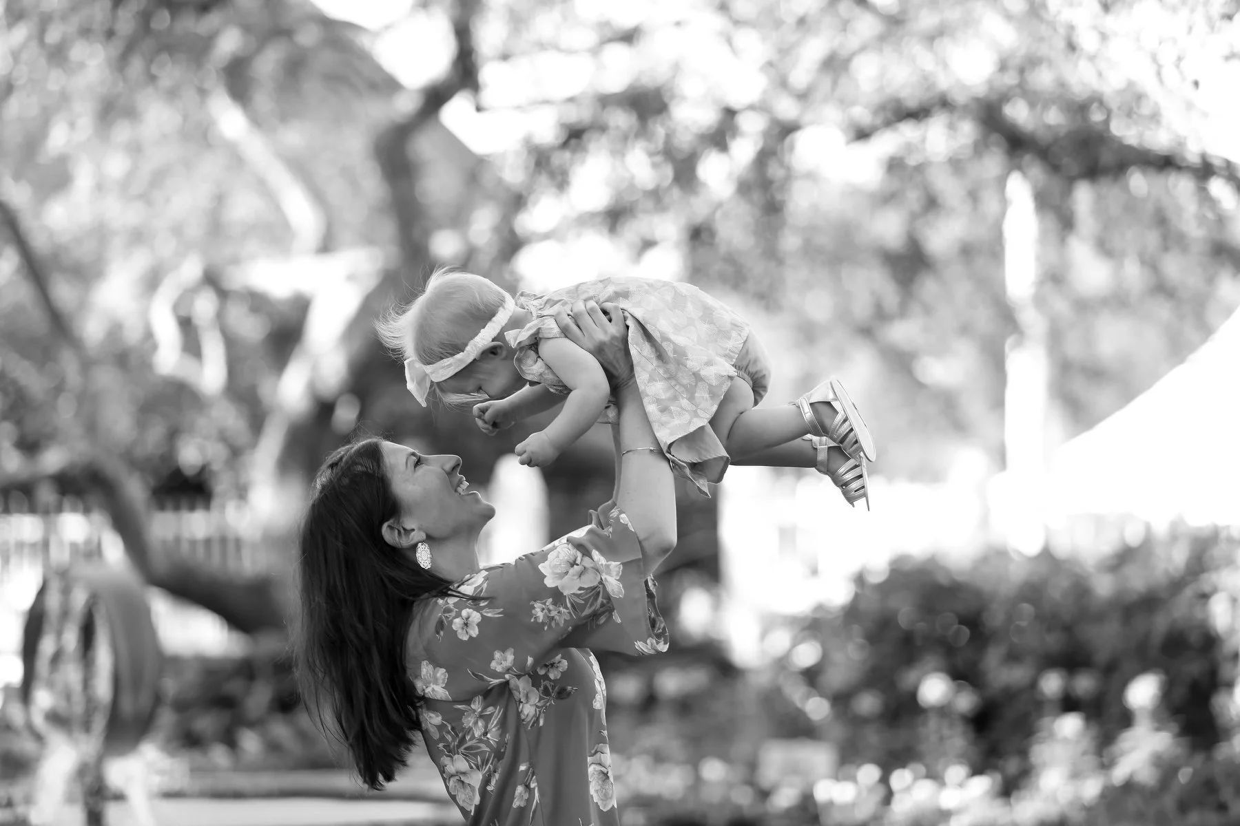A woman lifting a young girl in the air outdoors with trees in the background.