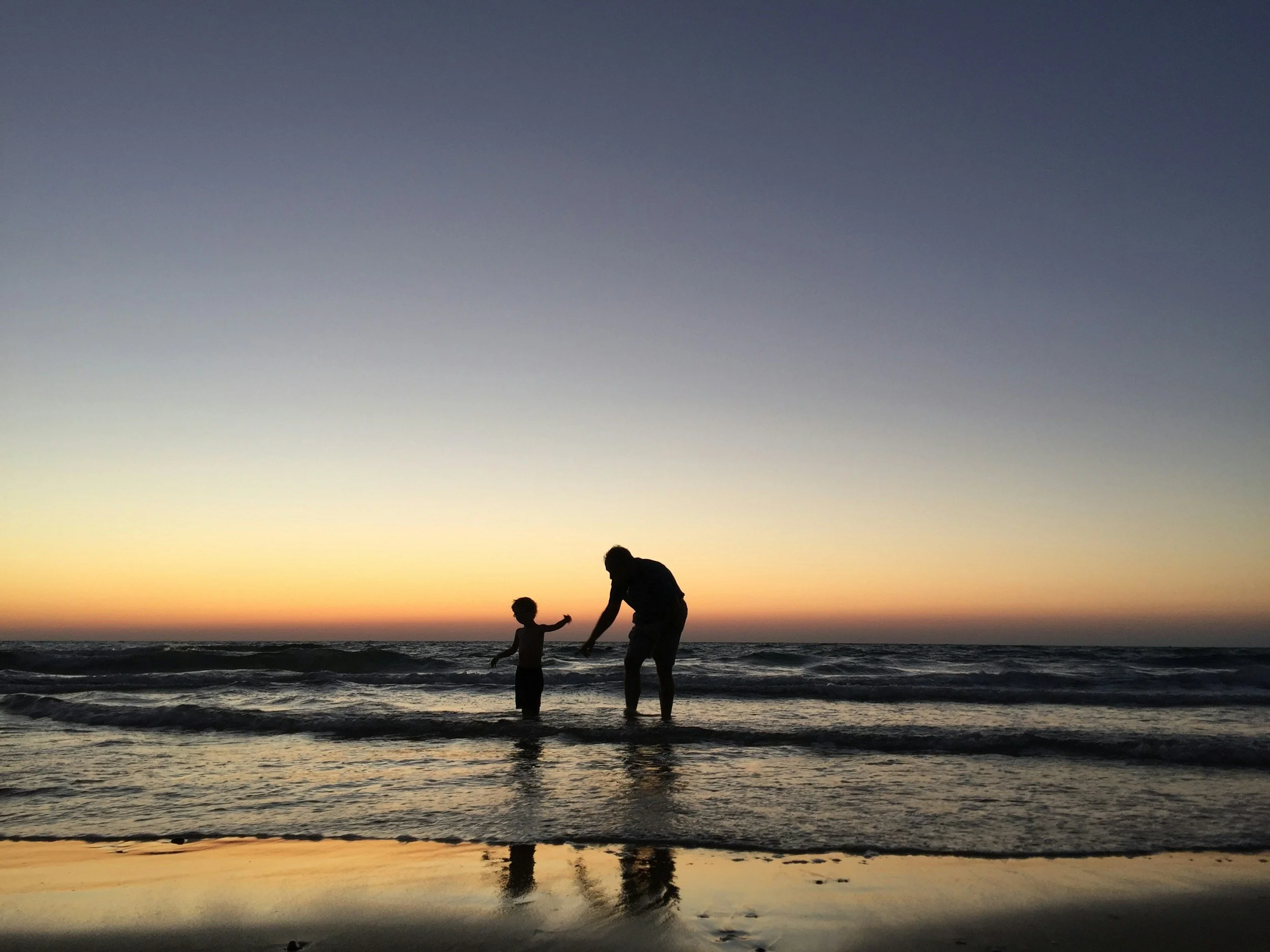 A silhouette of two people, likely a parent and child, holding hands and standing in shallow water on the beach during sunset or sunrise, with the sky transitioning from yellow near the horizon to dark blue at the top.