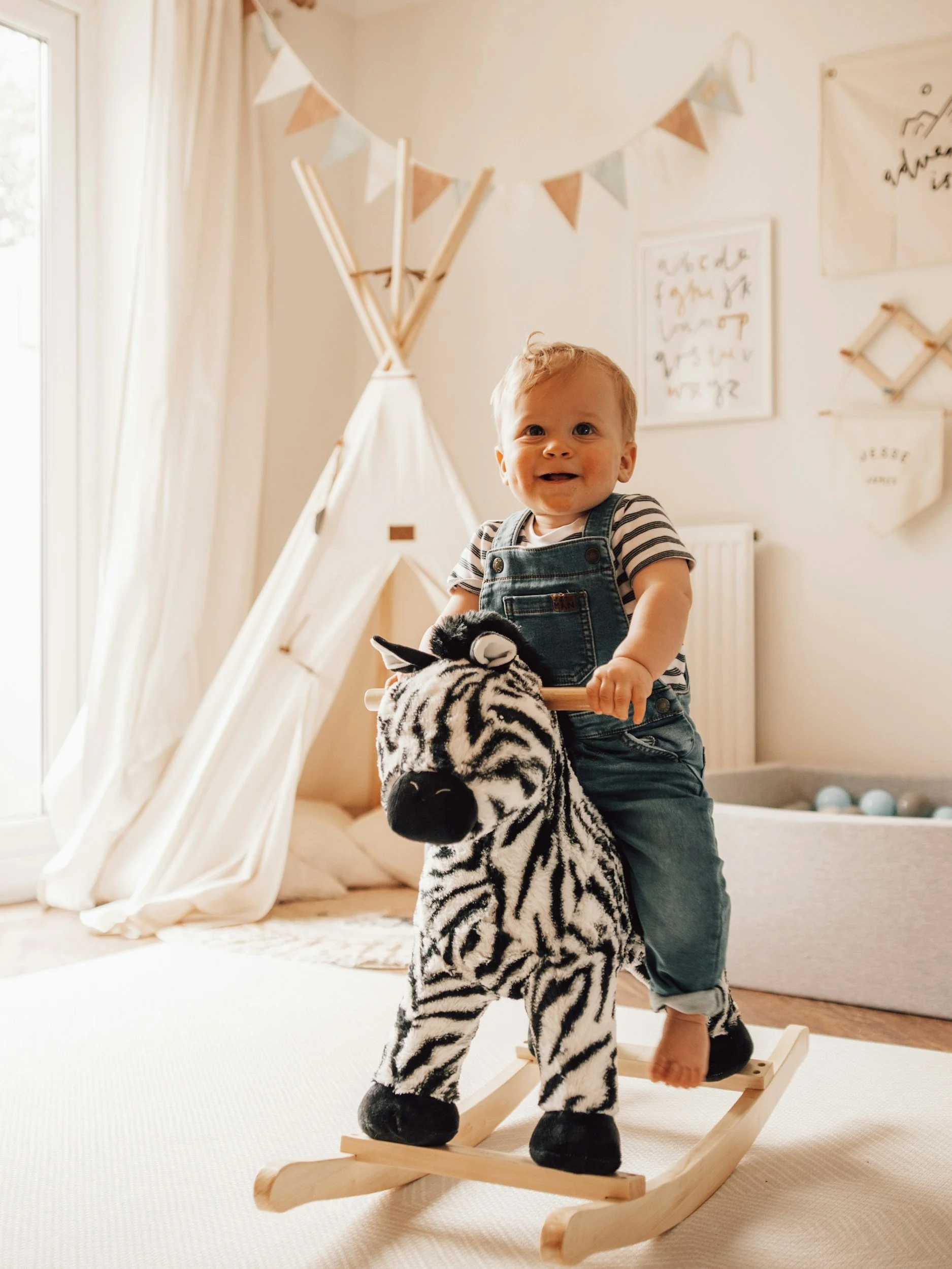 A young child riding a wooden rocking horse in a cozy, well-lit room decorated with a teepee, bunting, and wall art.