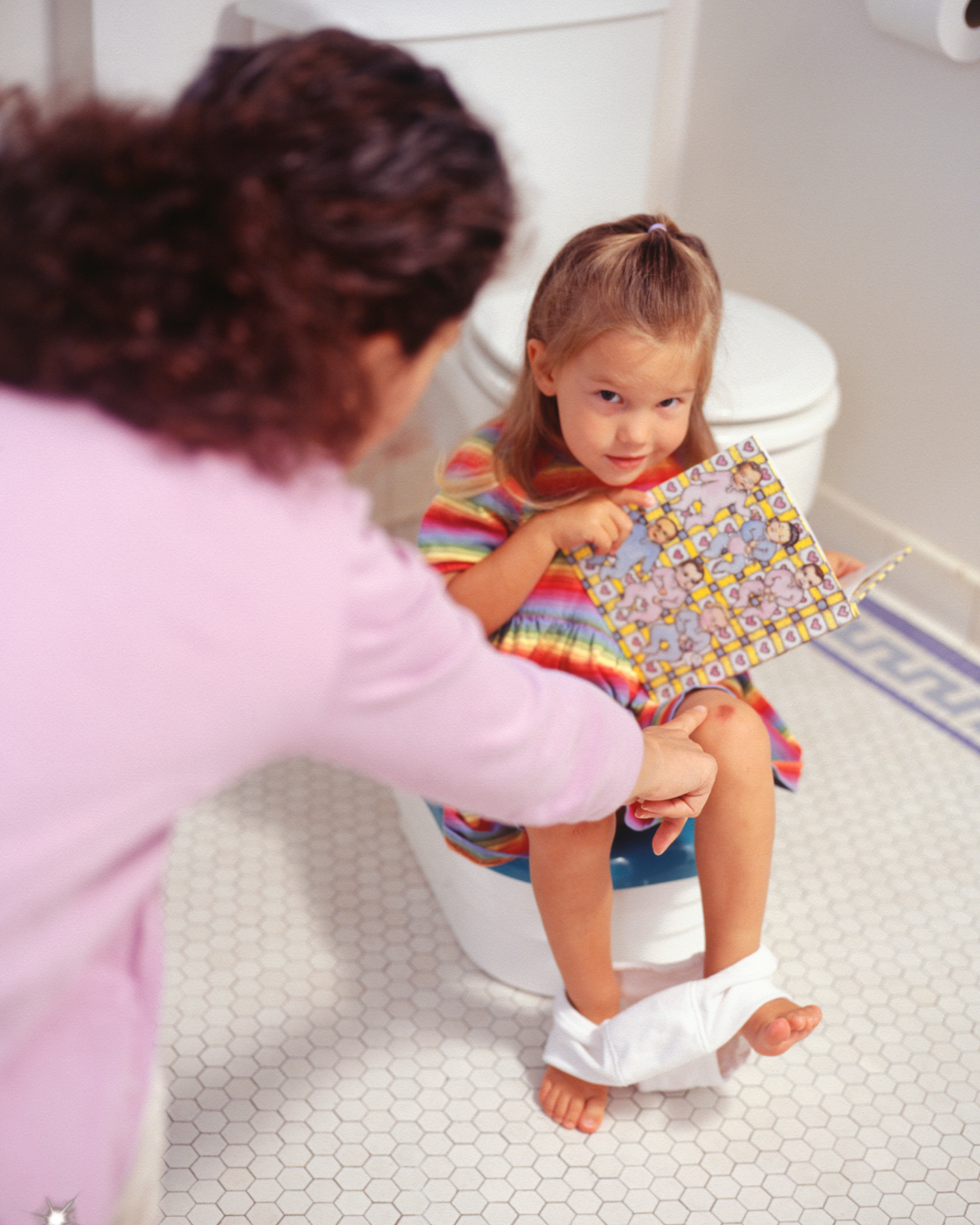 A young girl sitting on a toilet, holding a colorful book, being helped by a woman, possibly a caregiver or parent, in a bathroom with white tiles and a toilet. How to potty train Boston, MA