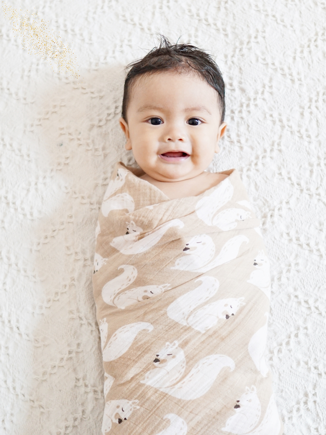 A smiling baby wrapped in a swan-patterned beige blanket, lying on a textured white surface.