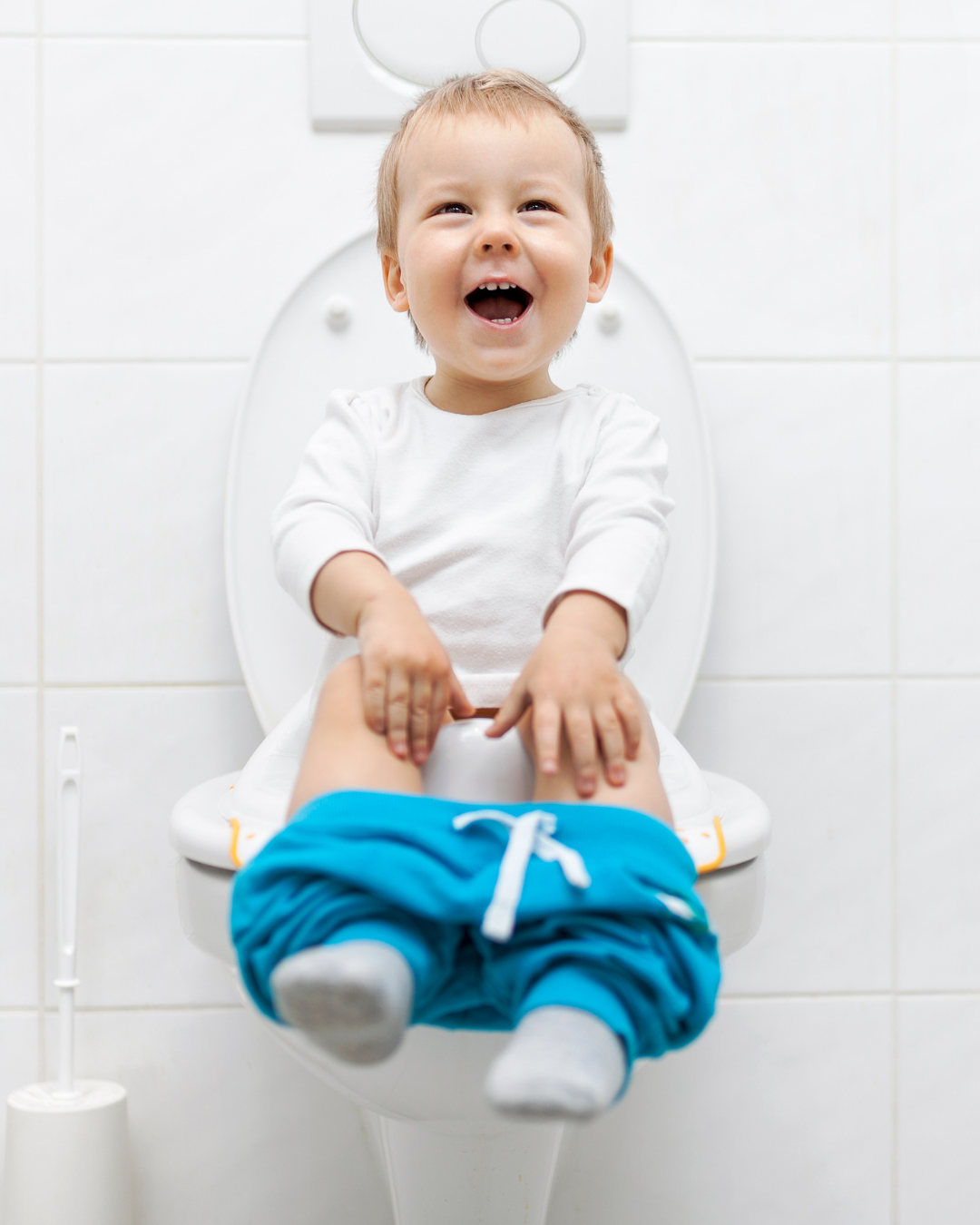 Child sitting on a toilet, smiling and playing, wearing a white shirt, blue pants, in a bathroom with white tiled walls.