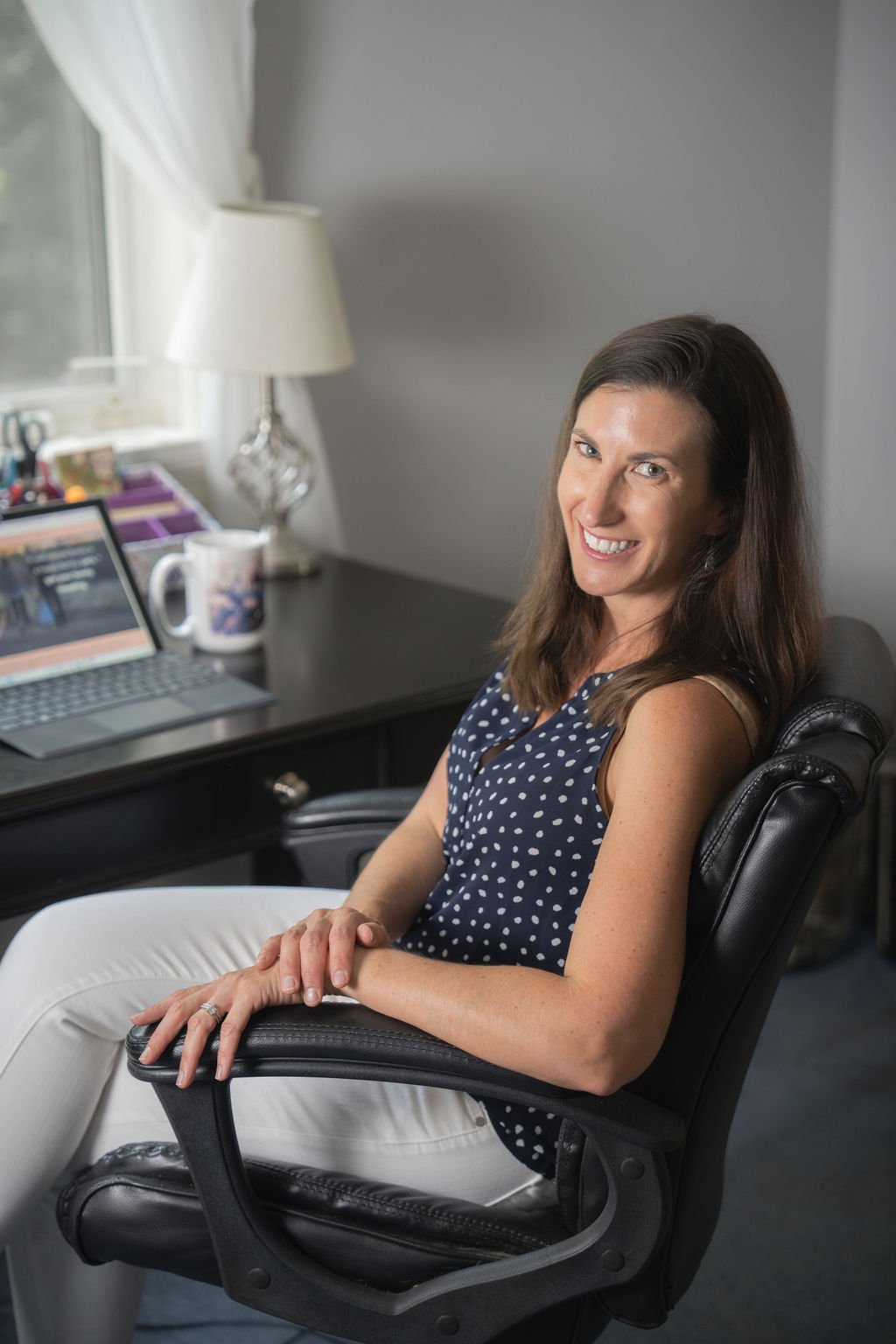 A woman sitting at a desk with a laptop, mug, and office supplies, smiling at the camera in a home office with a window and lamp in the background.