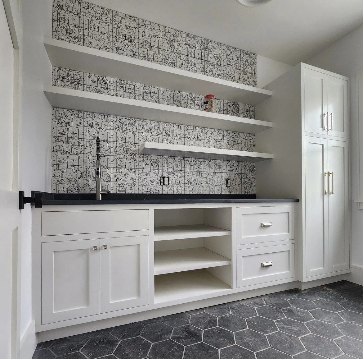 Kitchen with white cabinetry, black countertop, and patterned black-and-white wallpaper featuring cartoon characters. Open shelving with a red jar and black-tinted objects, hexagonal gray floor tiles, and a black kitchen faucet.