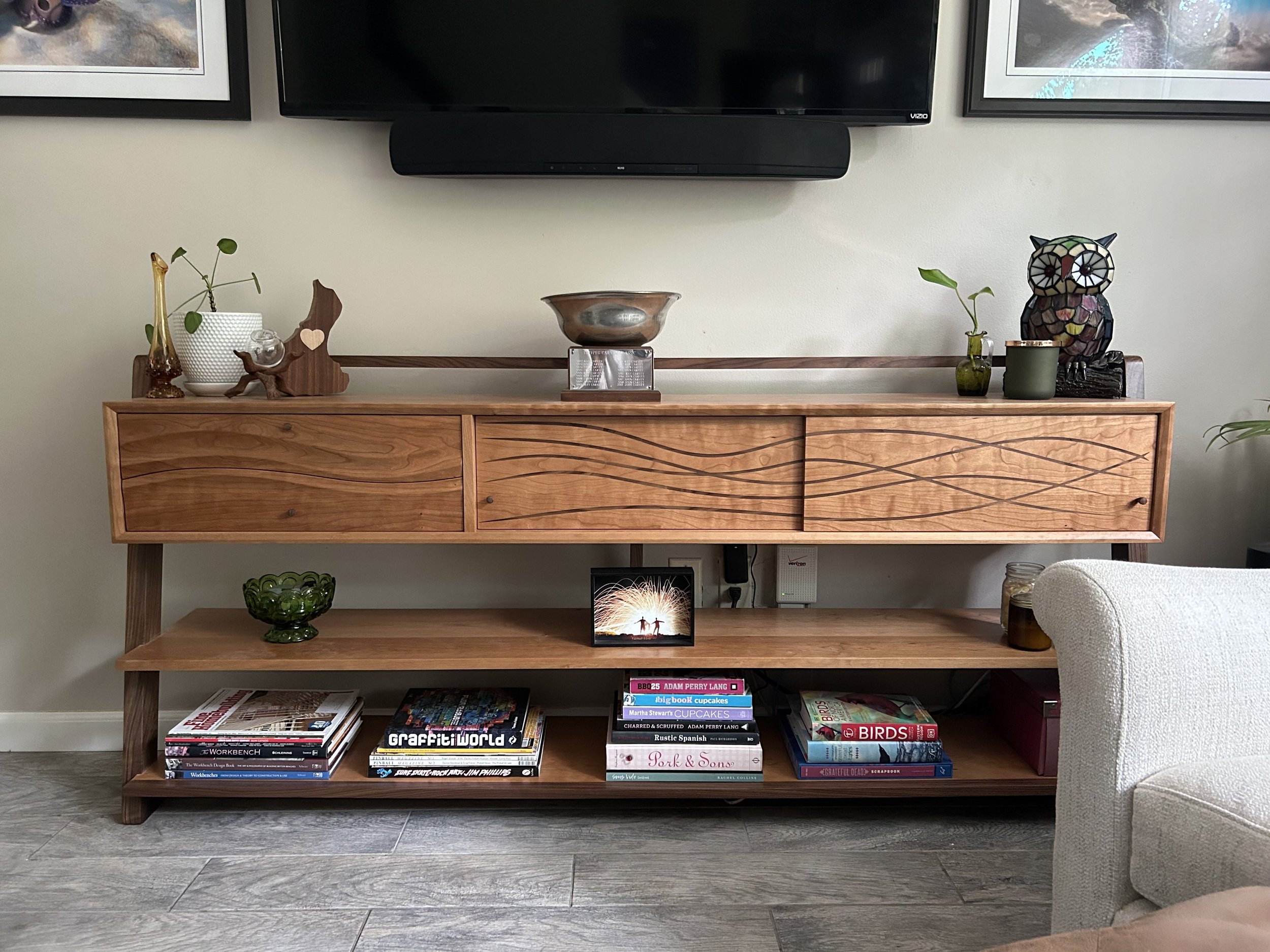 A wooden bookshelf with three levels holding various books, decorative items, and a small framed photograph. The top shelf has a small owl sculpture, a plant in a green vase, and a metal bowl. The middle shelf displays a stack of books, and a framed 