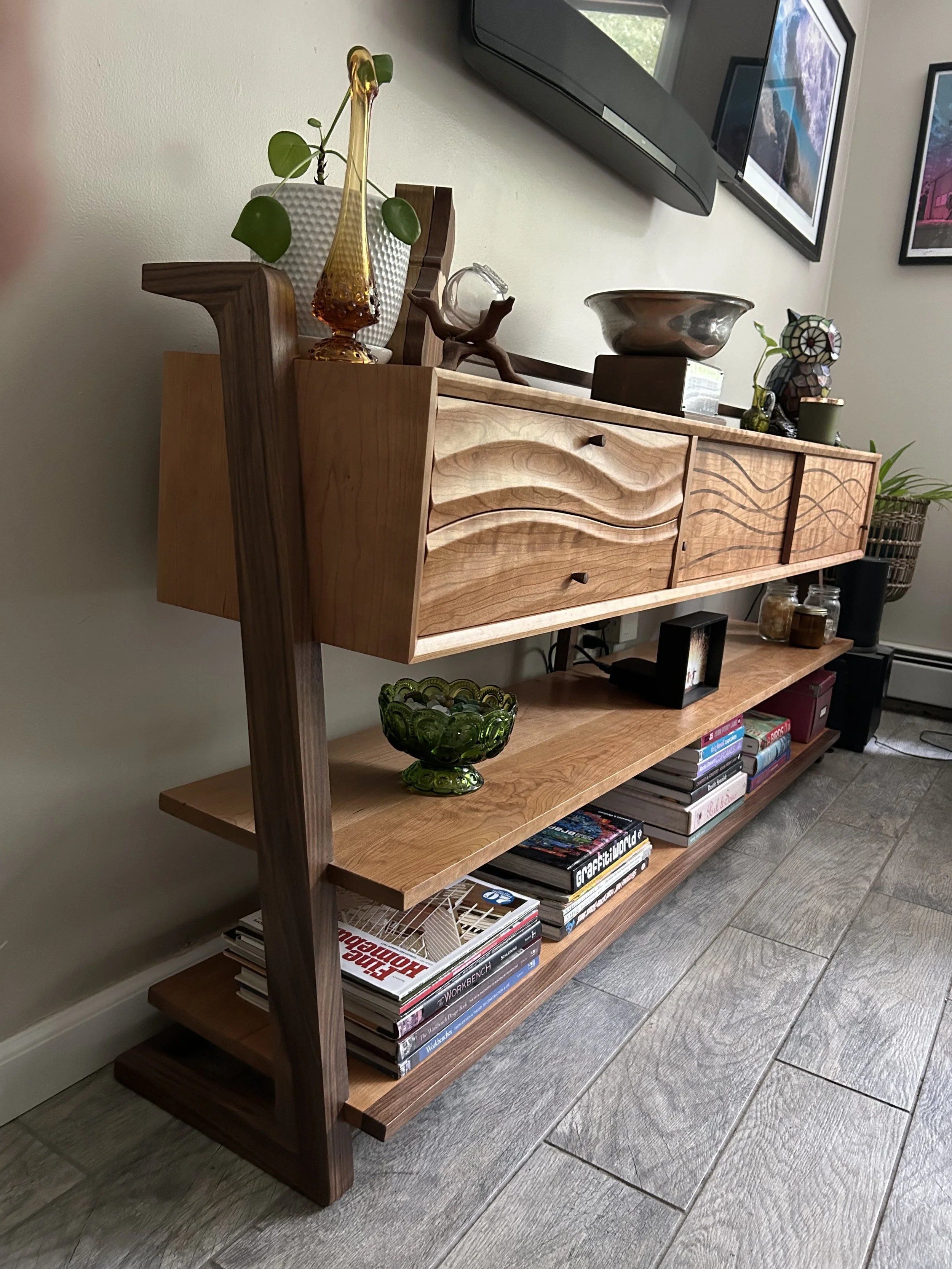 A wooden bookshelf with three shelves holding decorative objects, books, and magazines, with a wall-mounted TV and framed pictures in the background.