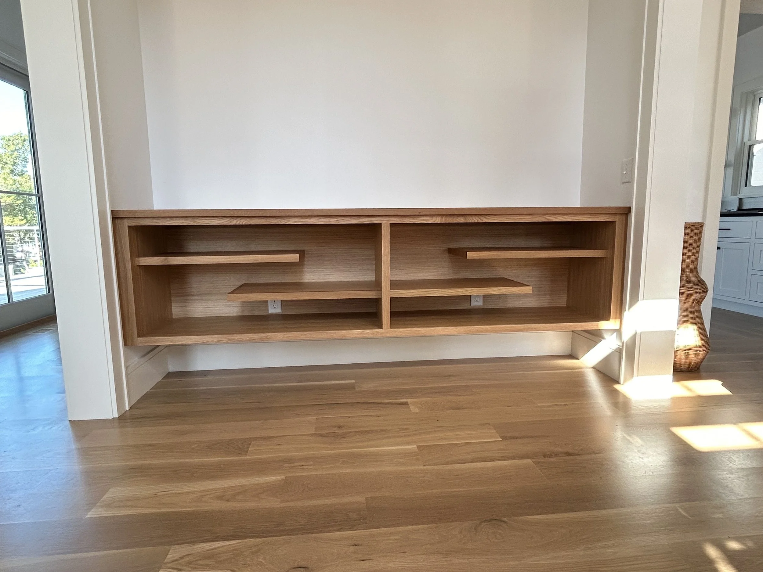 Empty wooden built-in bookshelf in a room with hardwood flooring and white walls, sunlight streaming in from the right side.