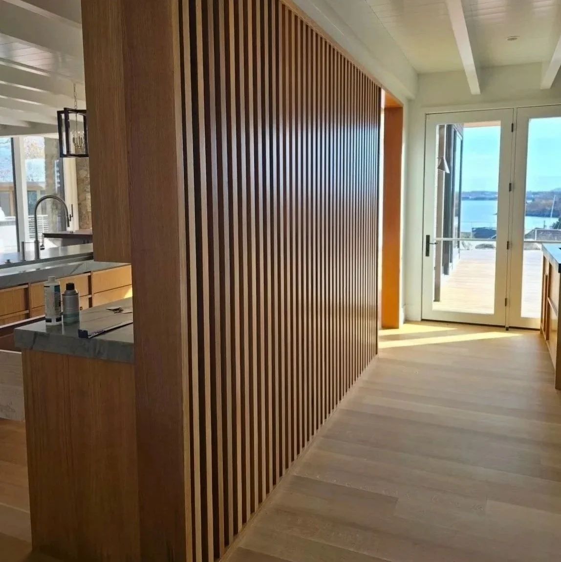 Interior view of a modern kitchen with wooden slat wall, white ceiling beams, and a glass door overlooking a water view.