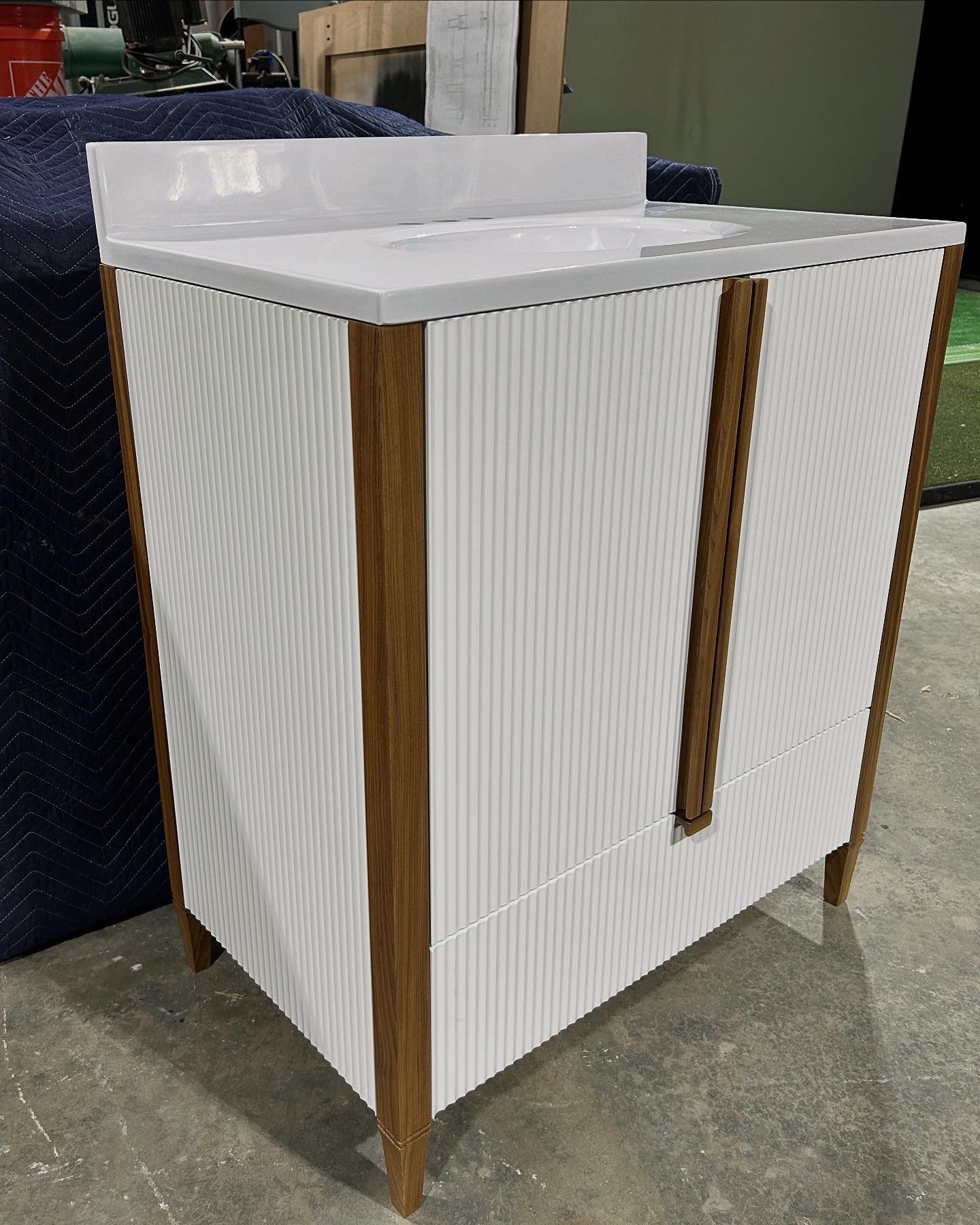 A white cabinet with wooden trim and fluted door panels stands on a concrete floor, with a countertop that appears to be a porcelain sink on top.