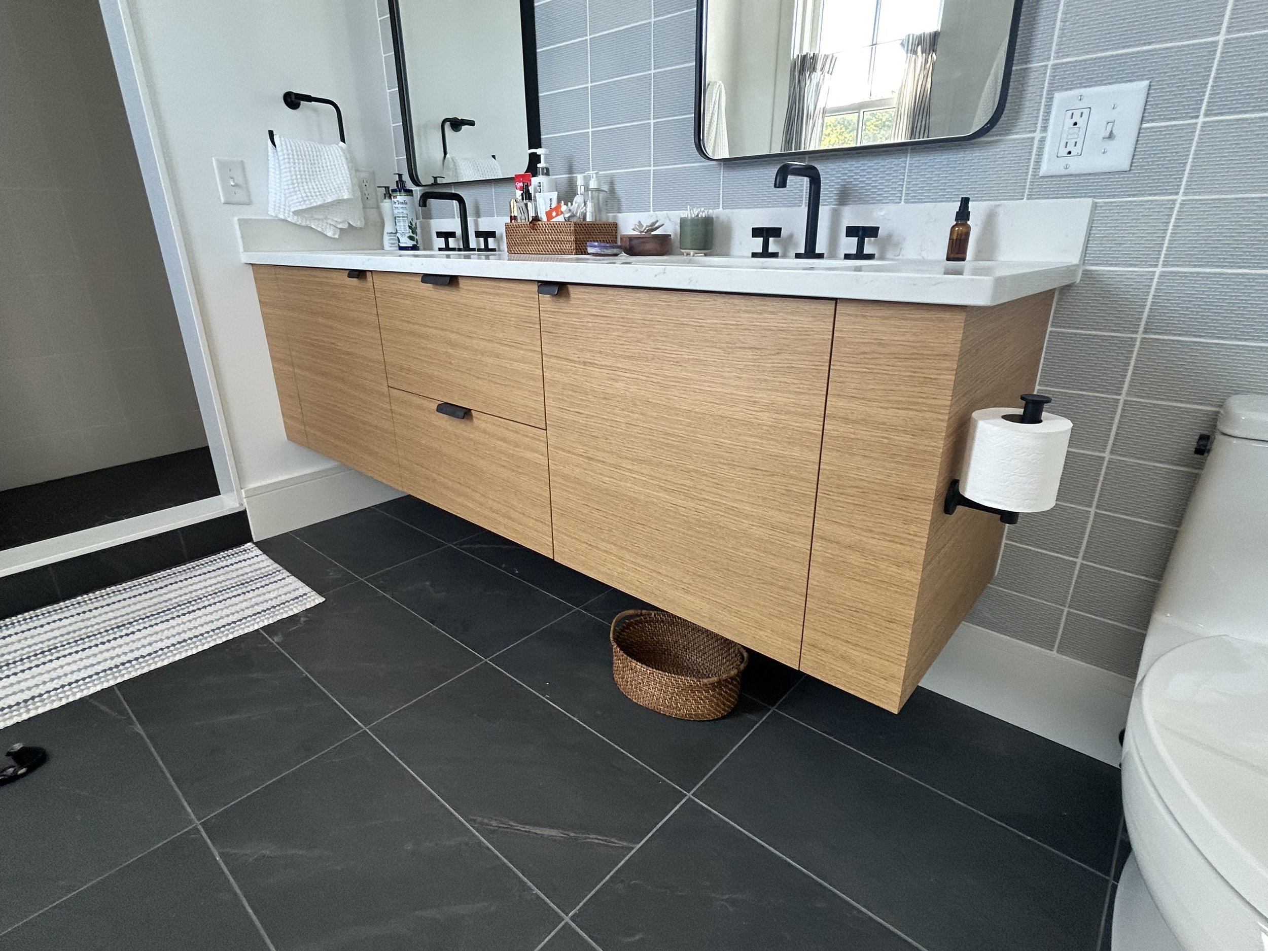 Modern bathroom with a light wood vanity, marble countertop, wall-mounted black faucet, and large mirror. Items on the counter include soap, lotion, and candles. A woven basket is on the floor next to the vanity. The bathroom has gray tiled walls and