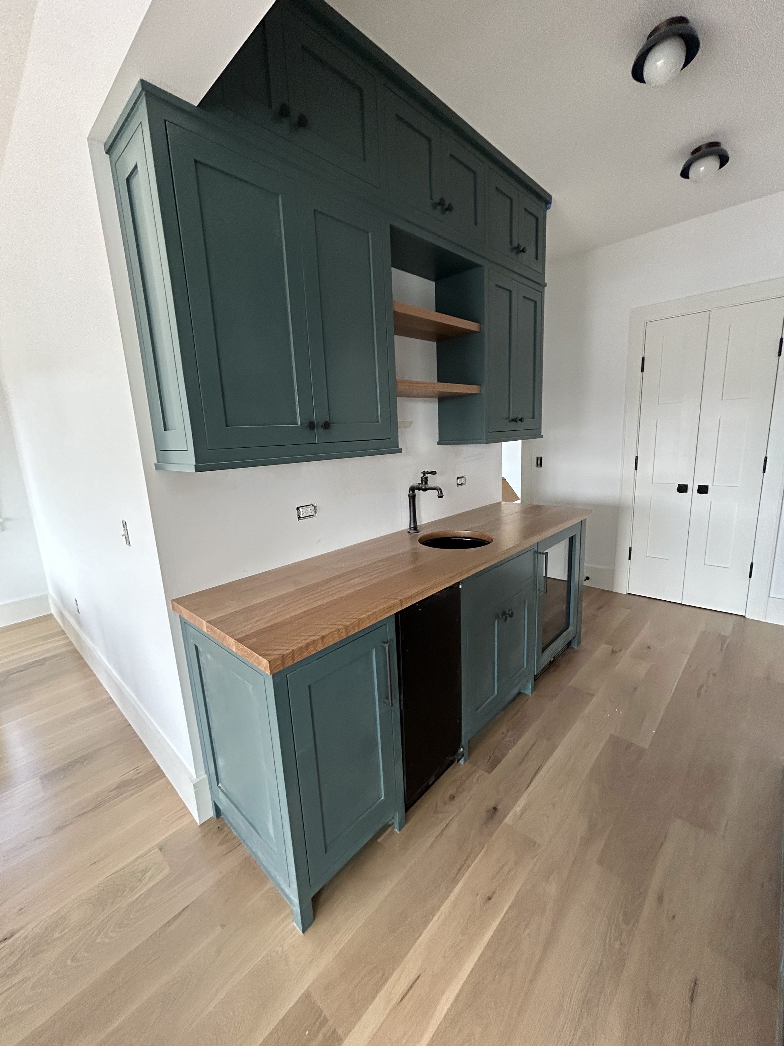 Photo of a kitchen area with teal cabinetry, open shelving, a wooden countertop, and a black sink. White walls and light wooden flooring. There are two black and white ceiling lights and a white door with black hinges.