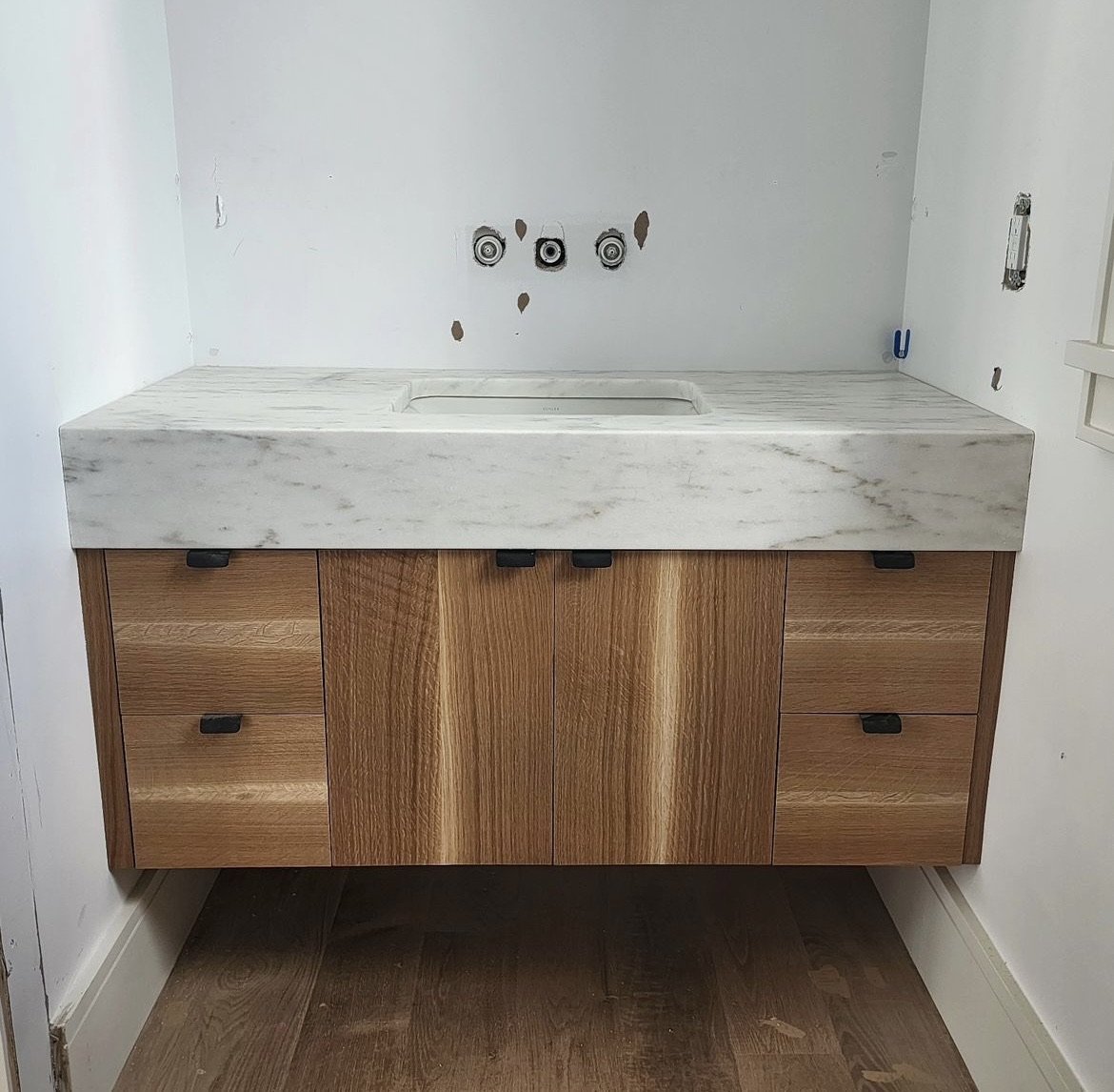 Bathroom vanity with a white marble countertop and wooden cabinets behind, with exposed plumbing and electrical fixtures on the wall.