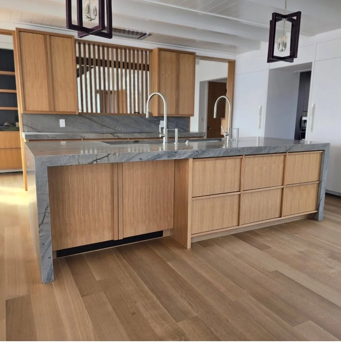 Modern kitchen with wooden cabinetry, marble countertops, and stainless steel sinks with high-arc faucets. Hardwood floors and contemporary pendant lighting are visible.