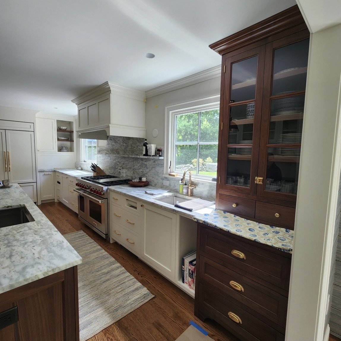 Kitchen with white cabinets, a granite countertop, a stainless steel oven with red knobs, a window above the sink, and a large dark wood cabinet with glass doors.