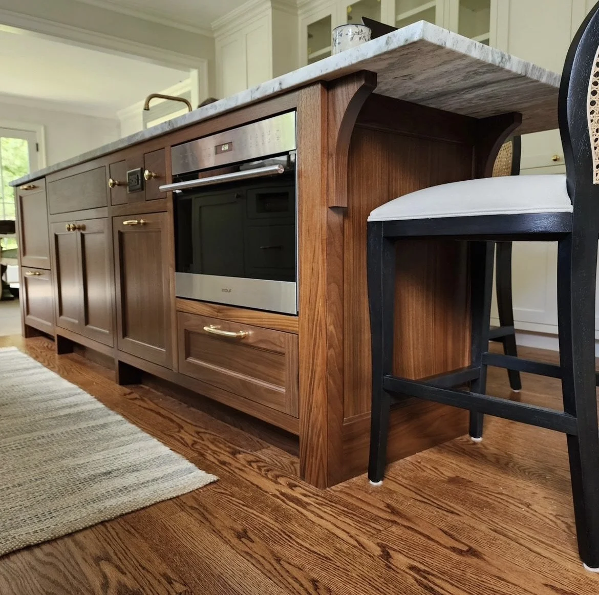 A kitchen island with wooden cabinets, a marble countertop, a built-in oven, and a black chair with a white cushioned seat beside it.