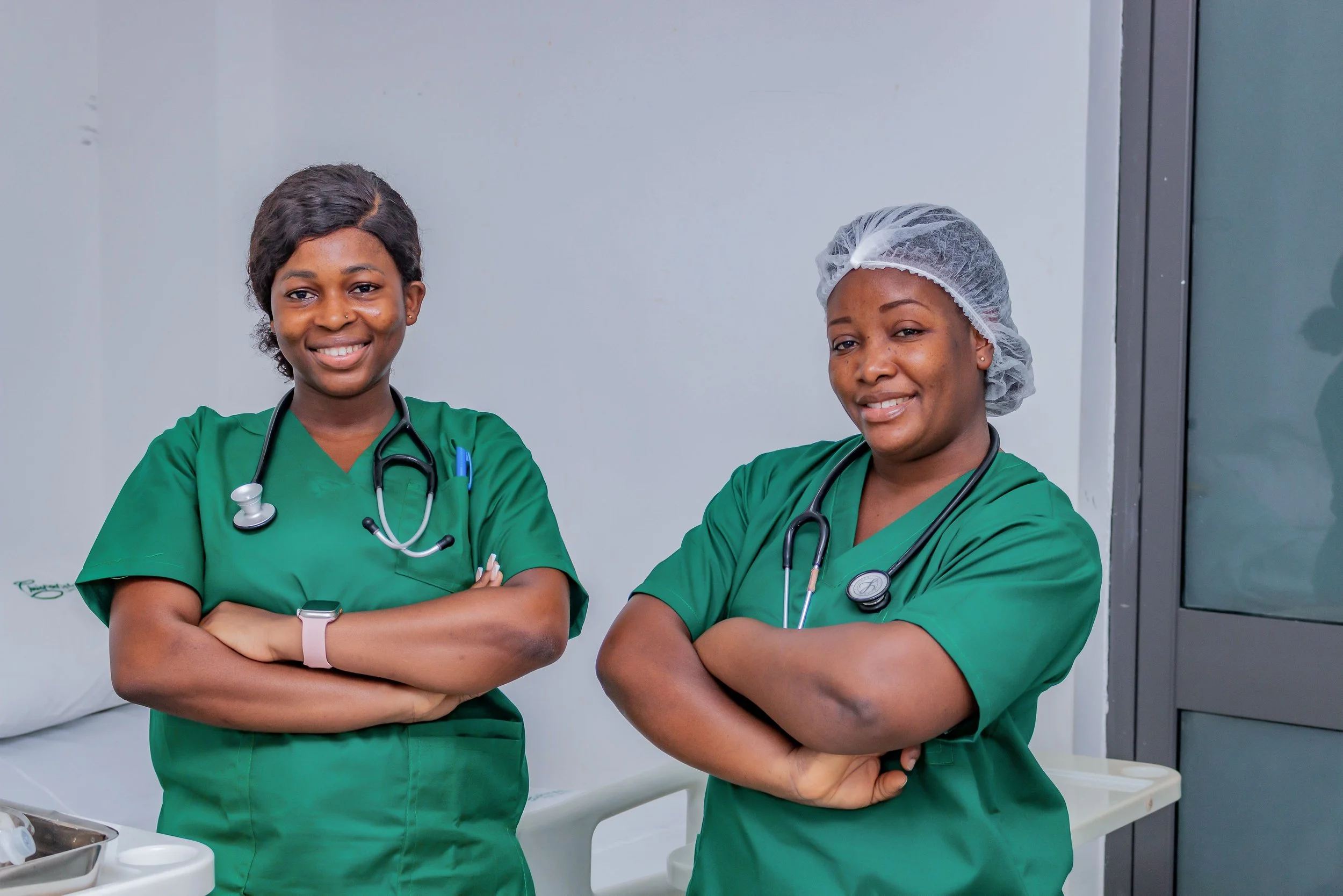 Two smiling nurses in green scrubs standing arms crossed in a hospital room