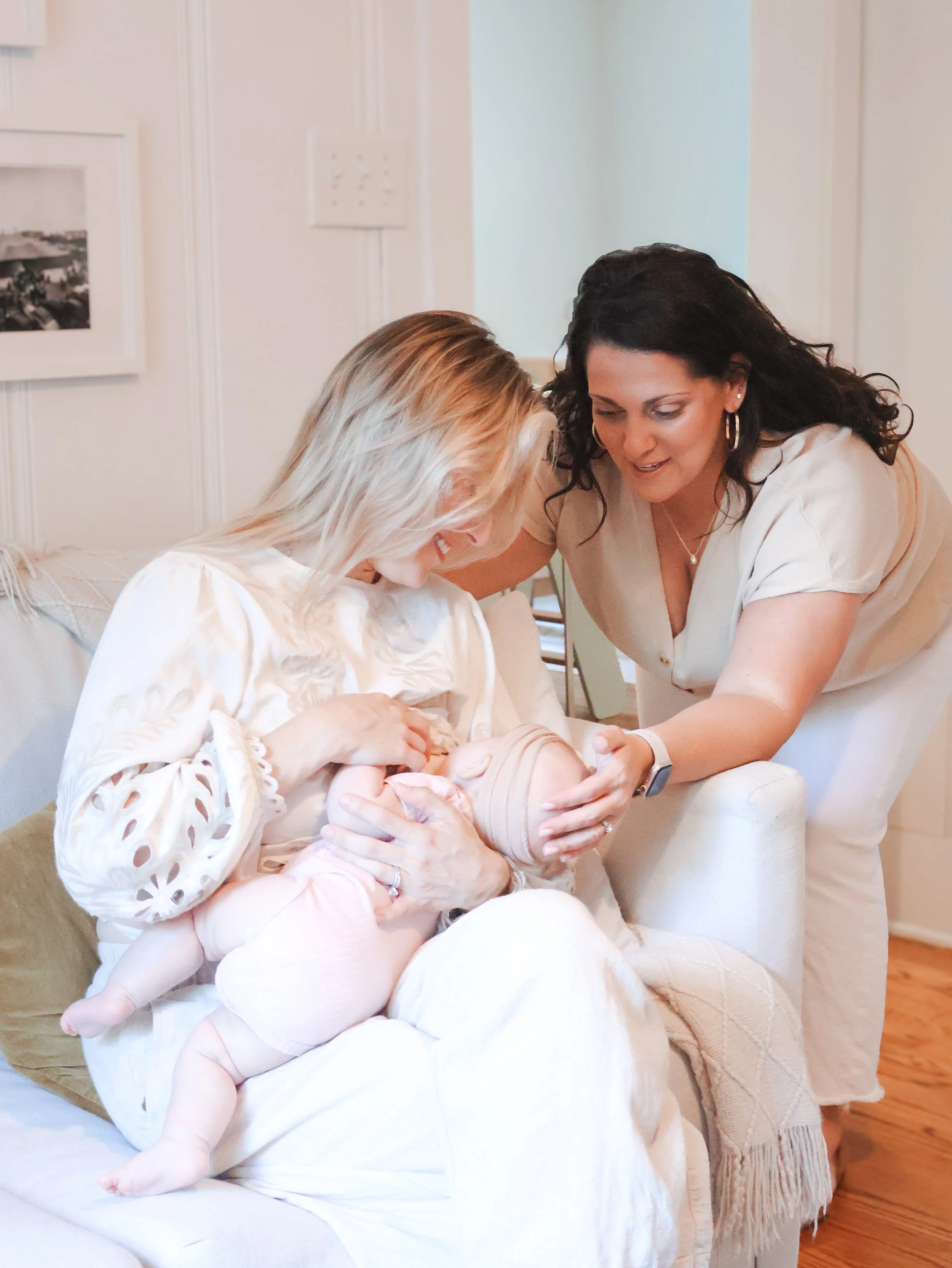 Two women, one with black hair and one with blonde hair, are smiling and interacting with a small baby in a cozy, well-lit room with white walls and wooden floors. The woman with black hair is wearing earrings and a light-colored top, while the blonde-haired woman is sitting on a couch, also smiling and engaged with the baby, who is lying on the woman's lap.