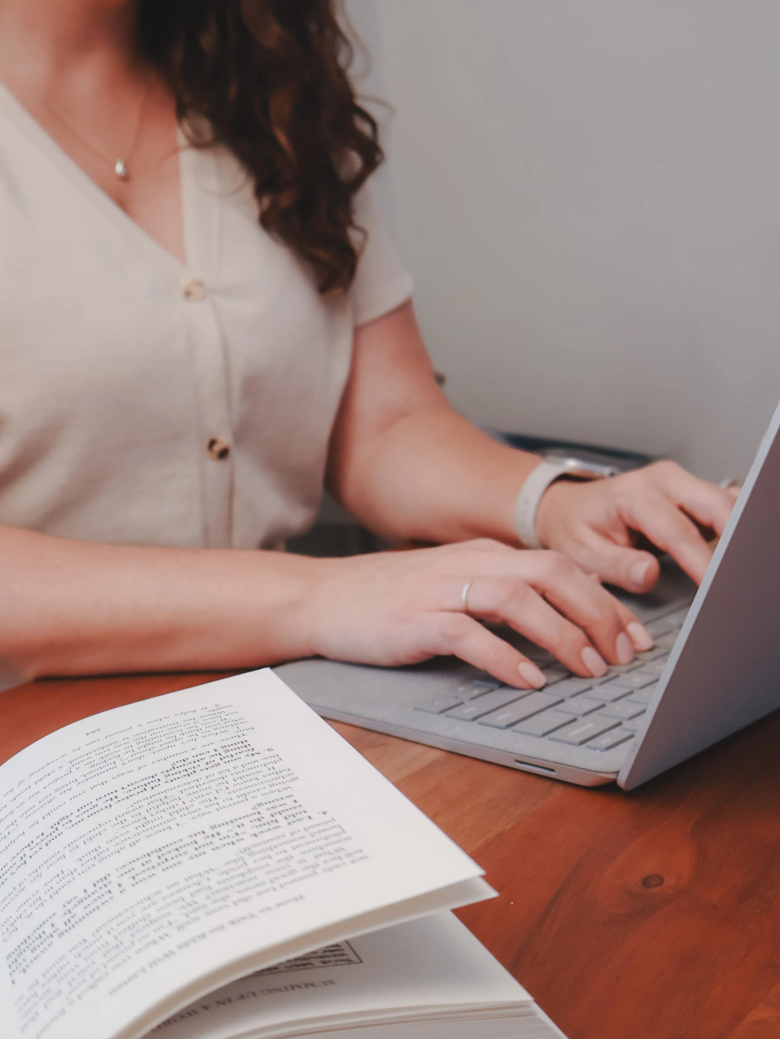 Person typing on laptop with open book and printed pages on wooden table.
