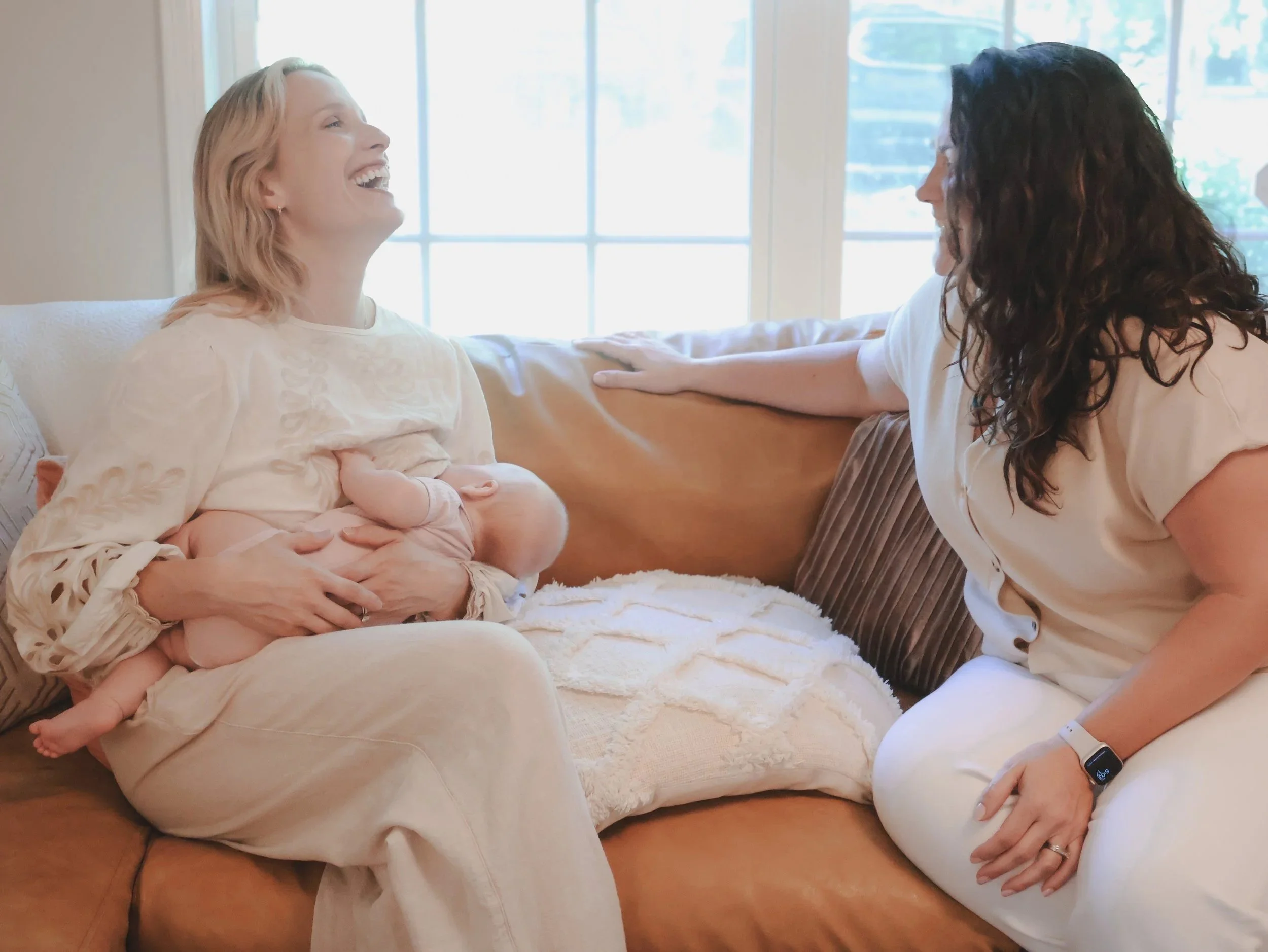 A woman with curly dark hair talking to a woman with short blonde hair who is holding a baby. The blonde woman is sitting on a beige couch near a window, and the dark-haired woman is leaning towards her with one hand on the couch armrest.