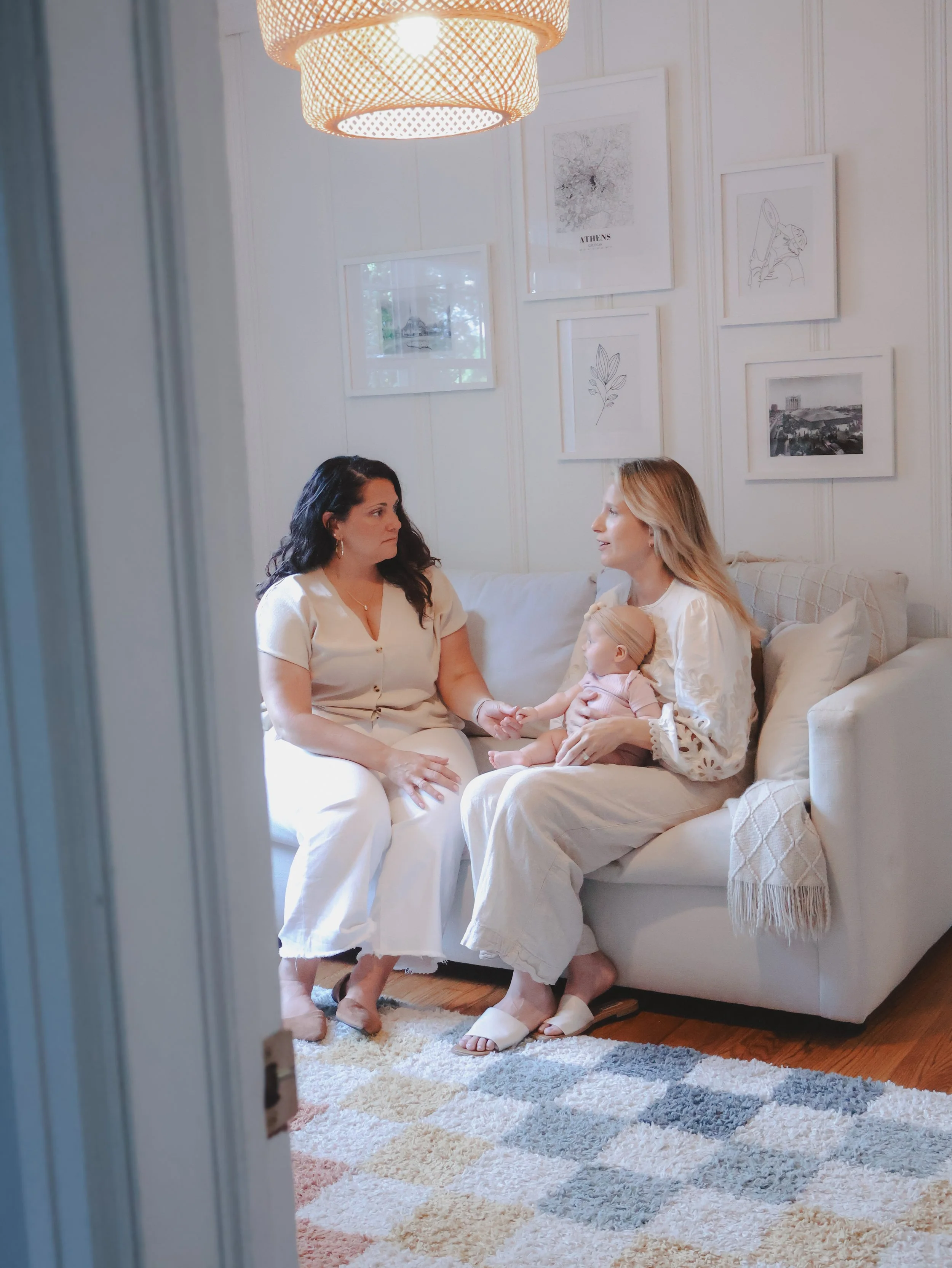 Two women and a baby sitting on a beige couch in a well-lit room. One woman has dark hair and is looking at the other woman, who has lighter hair and is holding a baby. The room has framed artwork on the white wall, a light-colored rug, and a woven pendant light hanging from the ceiling.