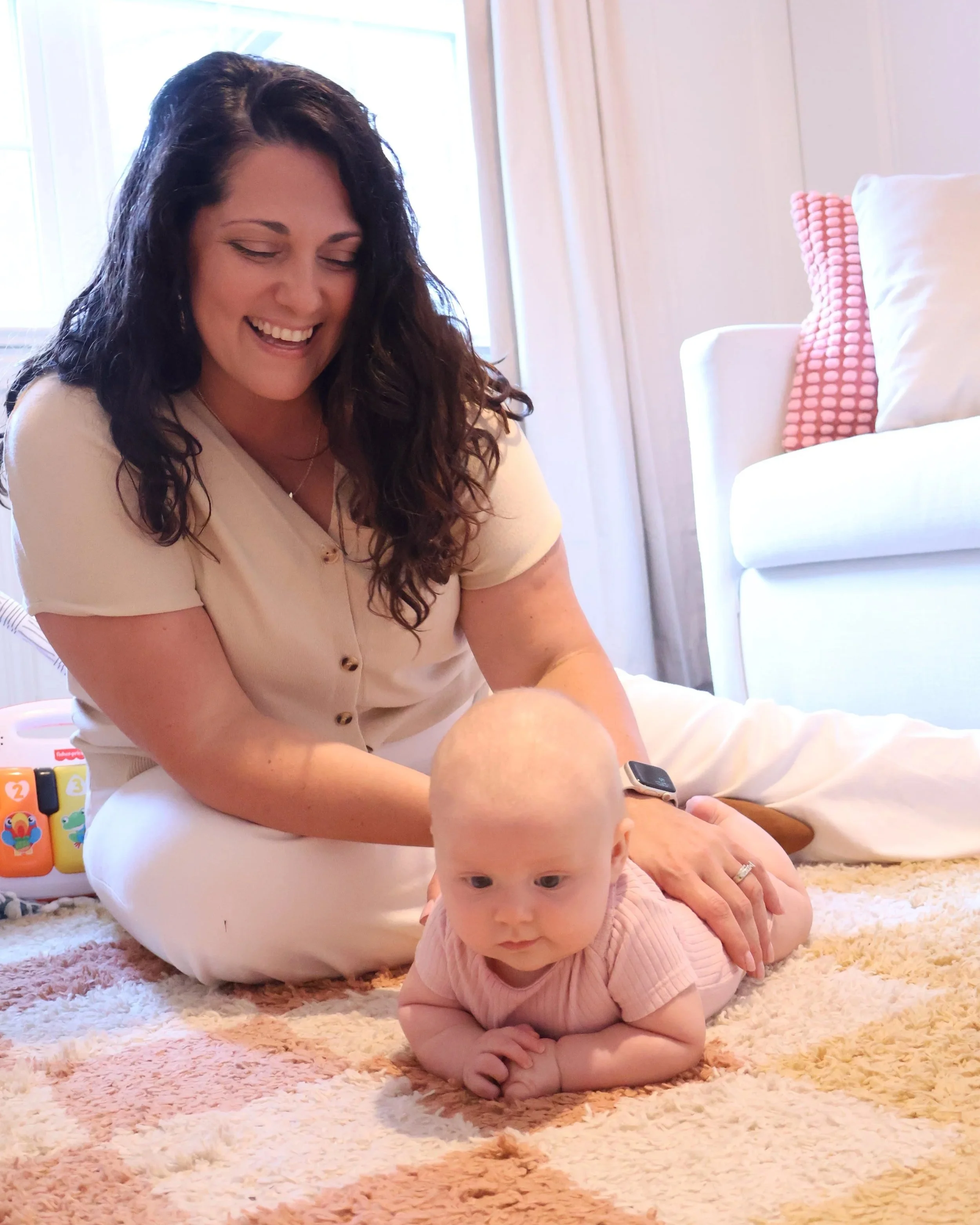 A woman with long dark wavy hair smiling and sitting on a beige shaggy rug with two children, a baby crawling in front and a toddler lying on the rug. There's natural light coming through a window in the background, and a white armchair with a pink textured pillow is visible.