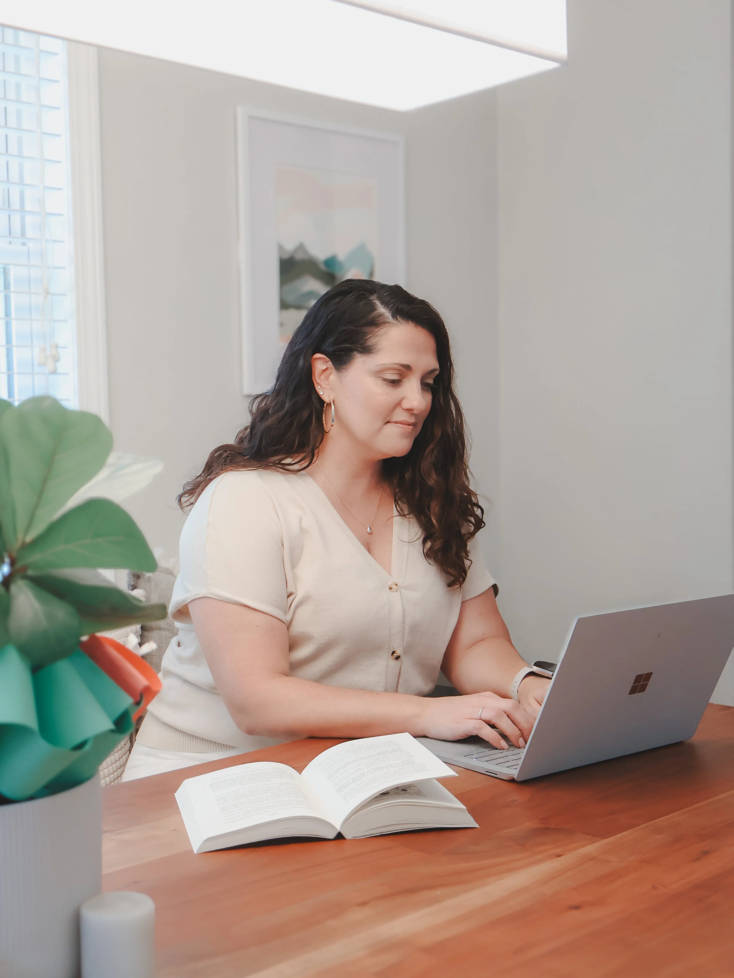 A woman with dark wavy hair, earrings, and a light-colored blouse sitting at a wooden table working on a silver laptop, with an open book in front of her and a potted plant on the side.