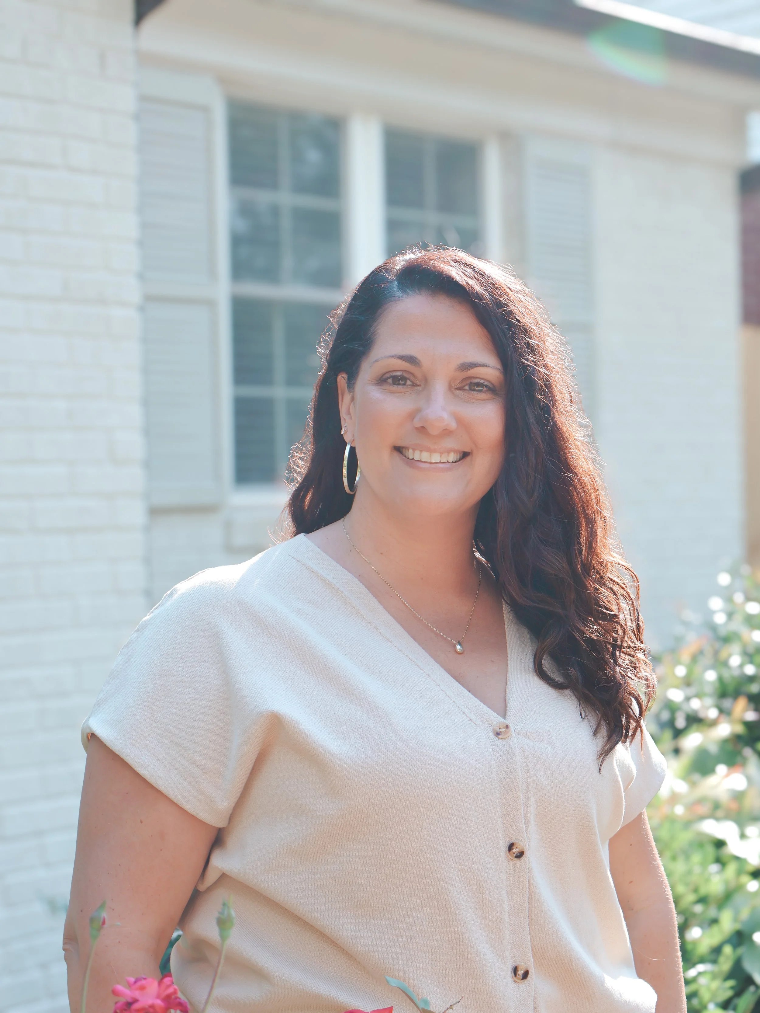 A woman with long dark hair, wearing hoop earrings and a light-colored, button-up top, standing outside in front of a house with large windows and a brick wall, smiling at the camera.
