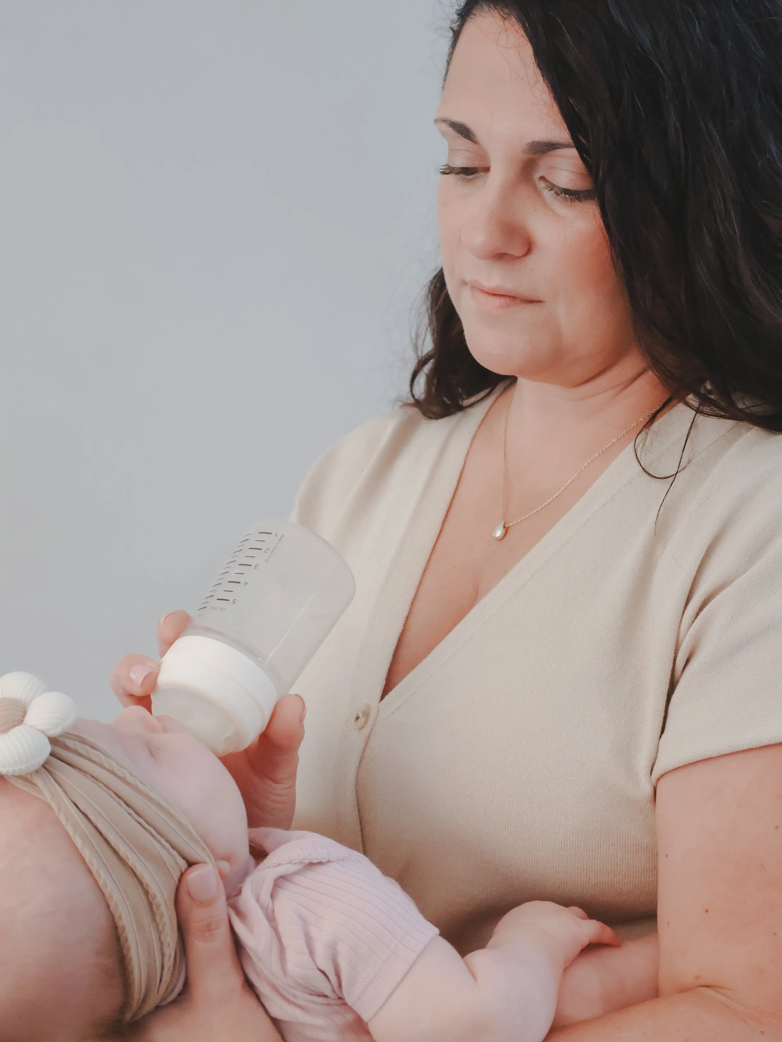 A woman with dark hair, wearing a beige top and a pearl necklace, holding a baby with light skin and wearing a pink and white striped outfit. The woman is feeding the baby from a bottle and looking down at the baby.