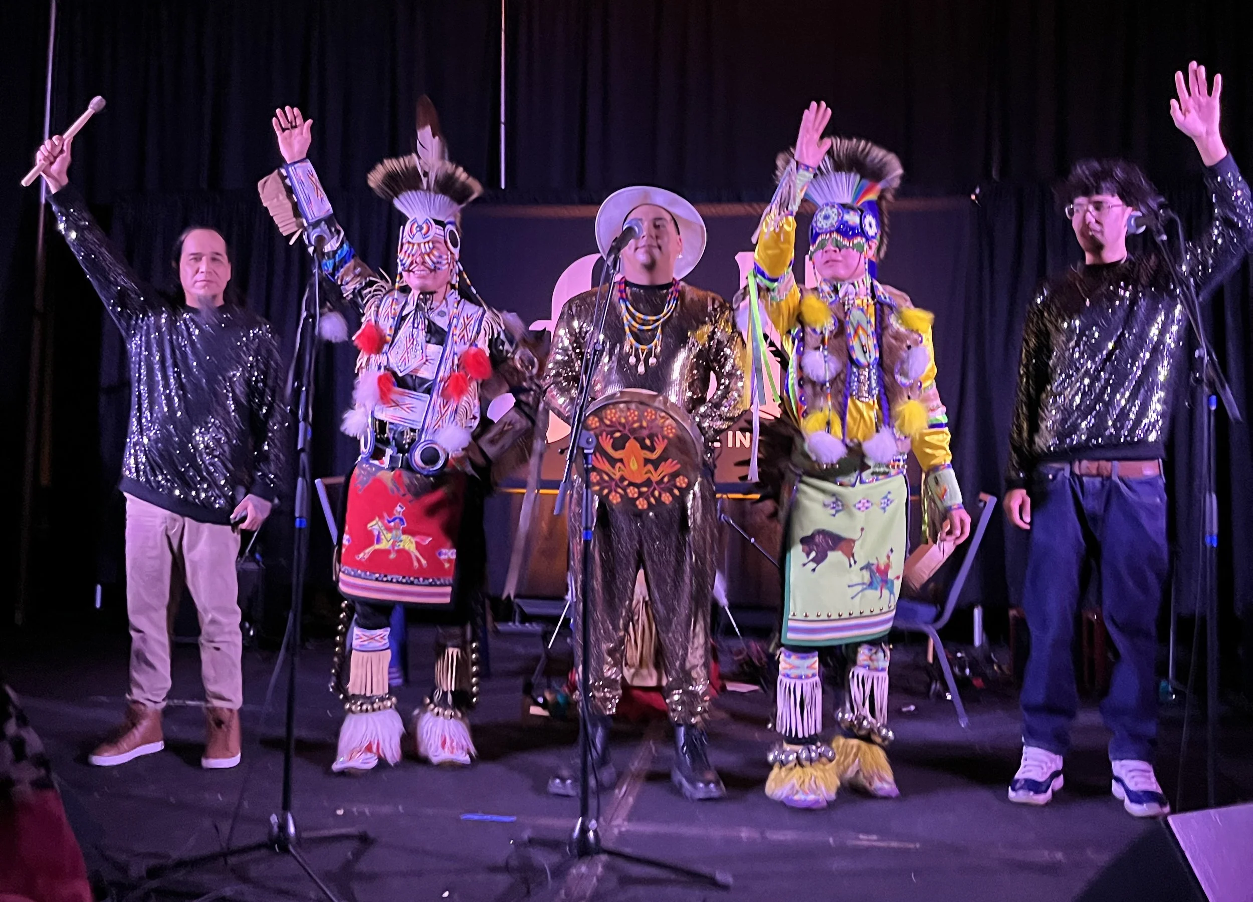 A photo of 5 Indigenous performers on stage, saluting the crowd. Some are wearing traditional Indigenous regalia.