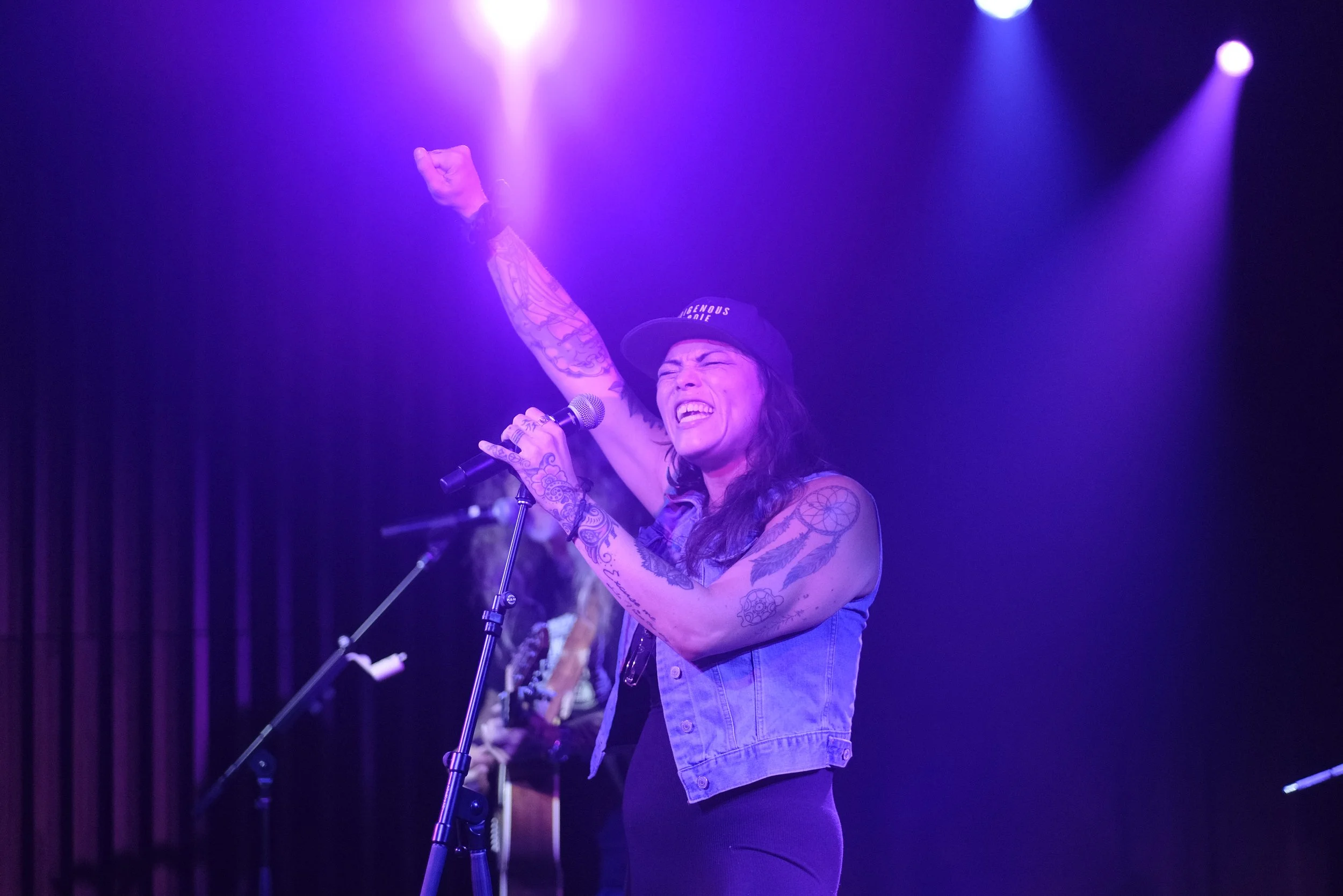 Six Nations artist Layla Staats is performing on stage, singing powerfully into the microphone with her right arm raised in the air and her fist clenched. It shows the strength and energy of Indigenous female artists.