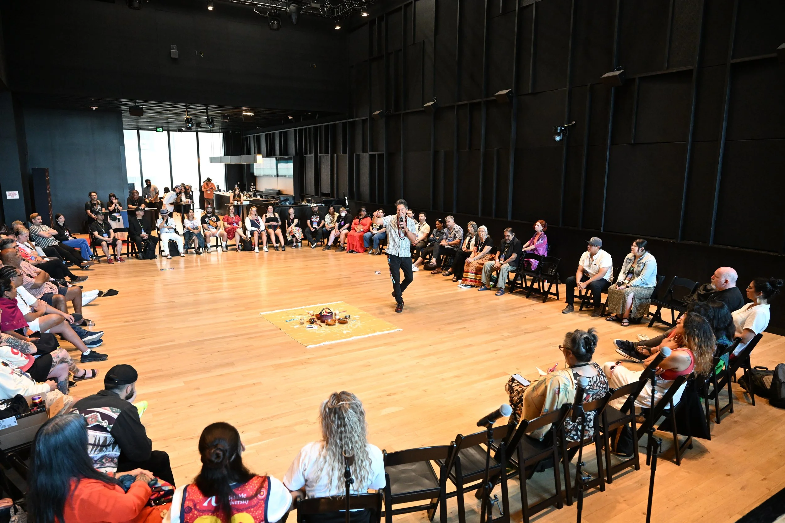 A photo of one of the circle sessions at IIMS. Raven is speaking into the microphone with 50-60 people in chairs sitting around him in a very big room at TD Music Hall. There are sacred objects and medicines on a blanket in the middle of the circle.