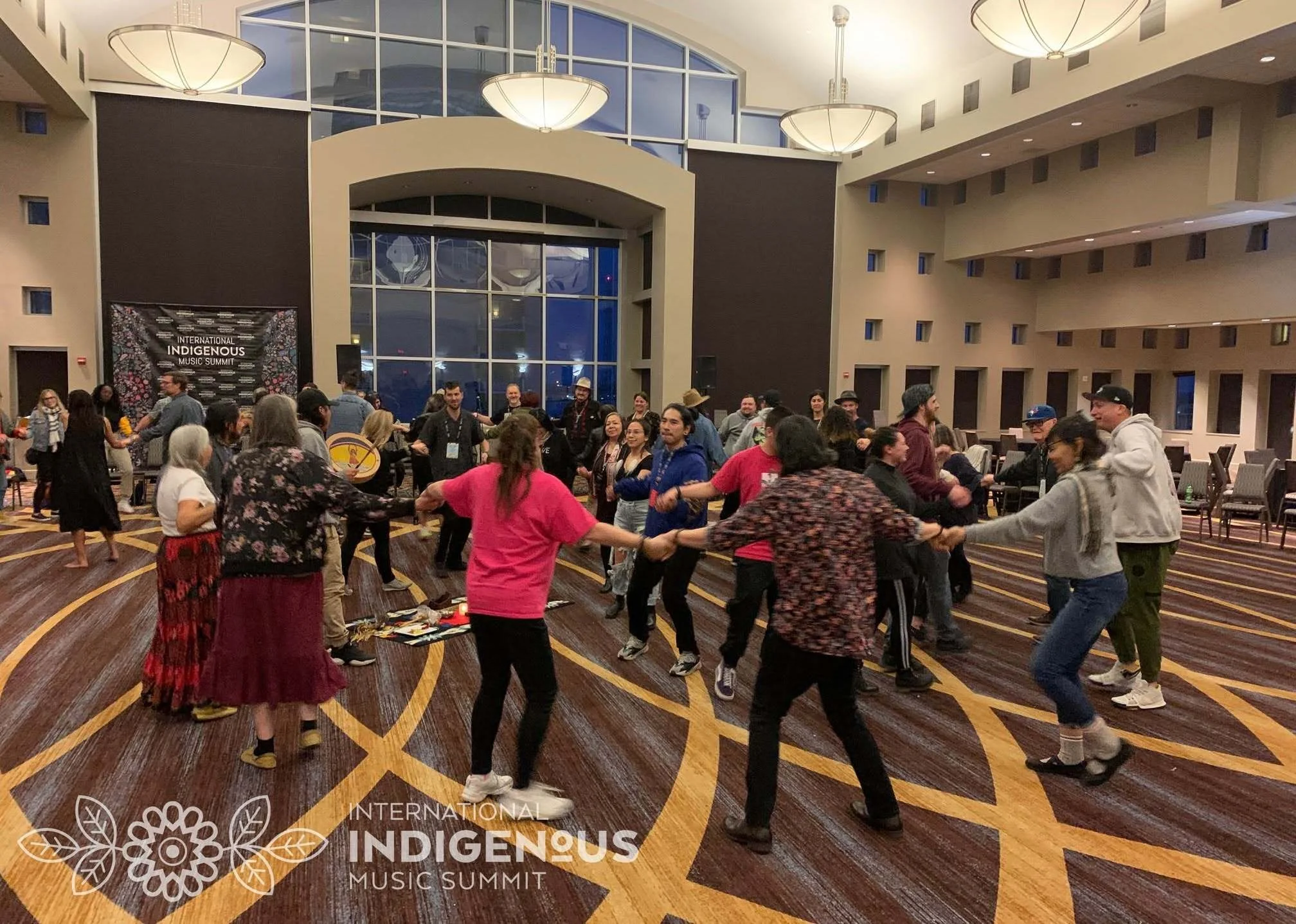 A photo of a group of people in a large room at a hotel, hand in hand, dancing while someone plays a drum.