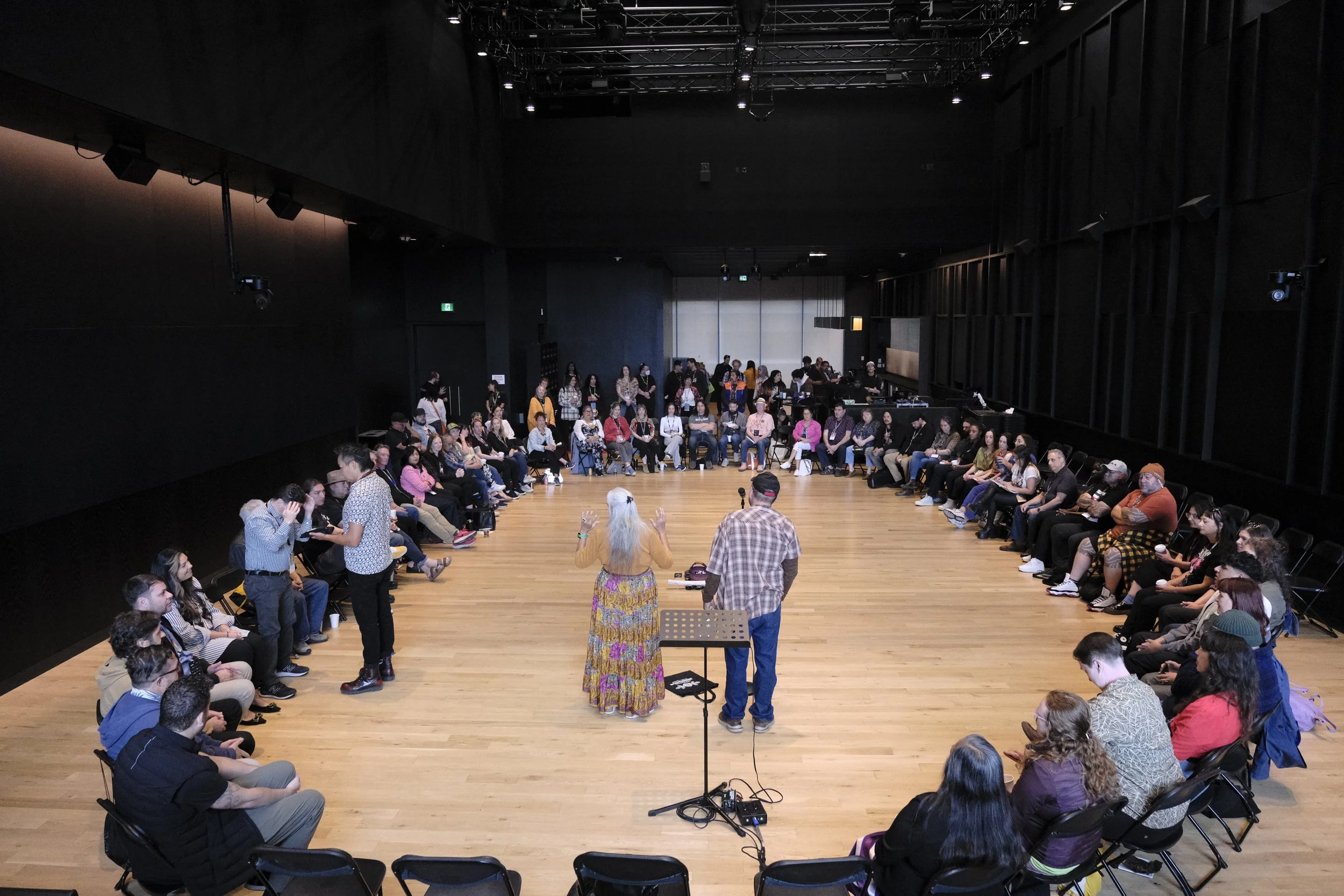 Photo of a ceremony and keynote speech. Elders are speaking to a crowd in a large circle of 50-70 people. Some participants are being smudged.