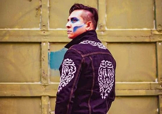 Photo of Jorge Requena Ramos with his back to the camera, looking over his shoulder into the distance, with drastic blue & white face paint on his cheeks, mouth and eyes.