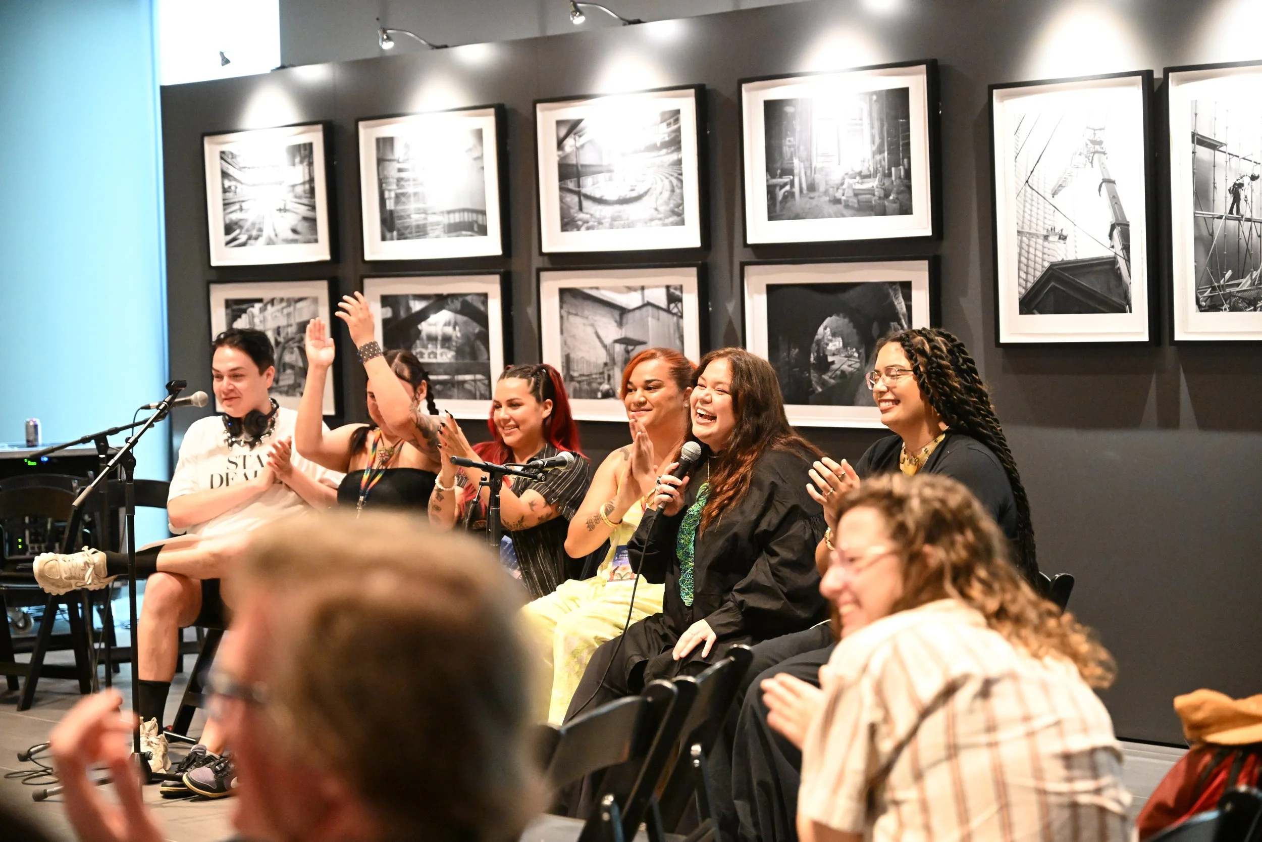 A photo from one of the IIMS's many industry panels shows a row of 6 panel speakers sitting beside each other. There is laughter and clapping. In the foreground, audience members are also clapping.
