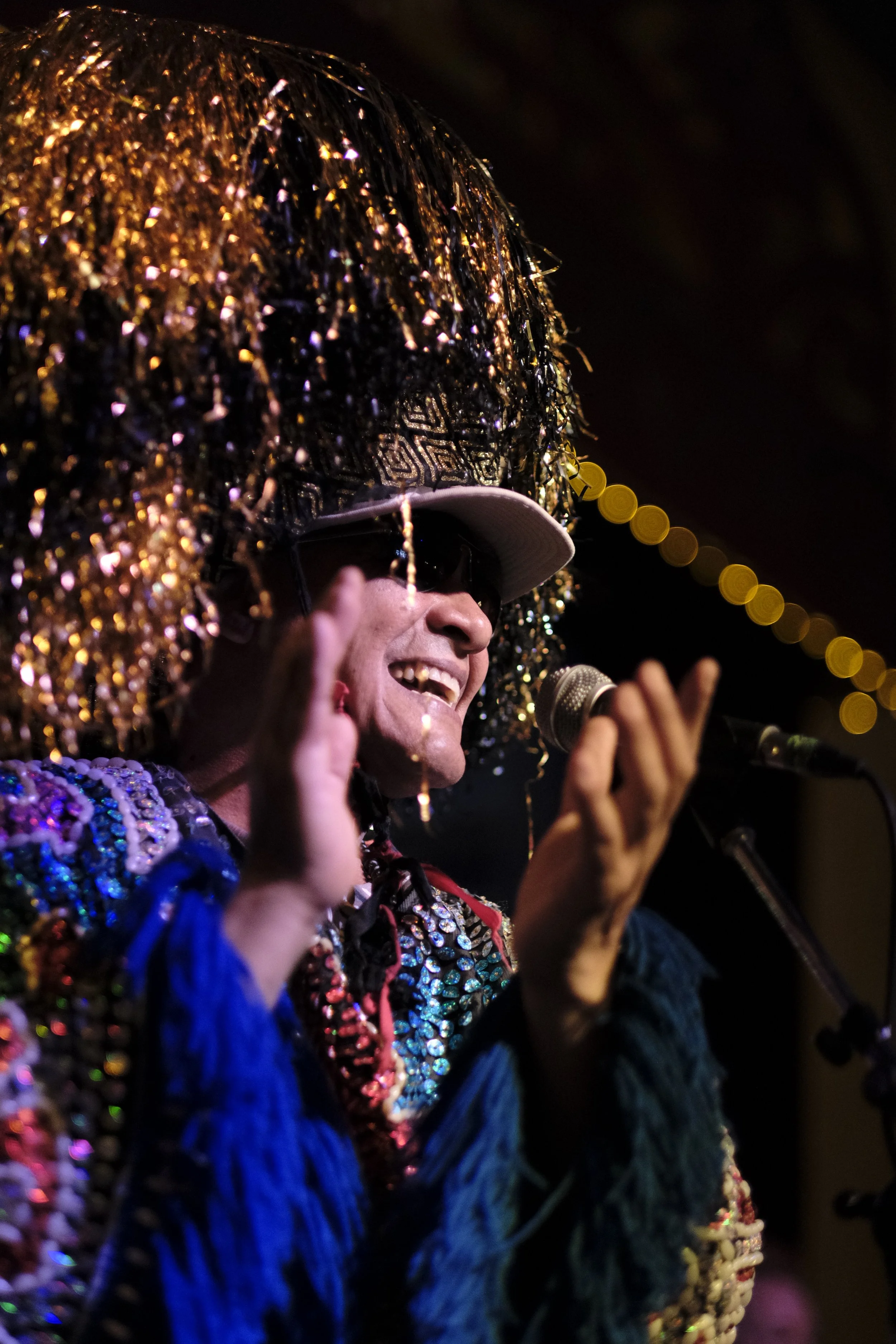 A performer in colourful, sparkling traditional regalia is on stage, smiling and clapping.