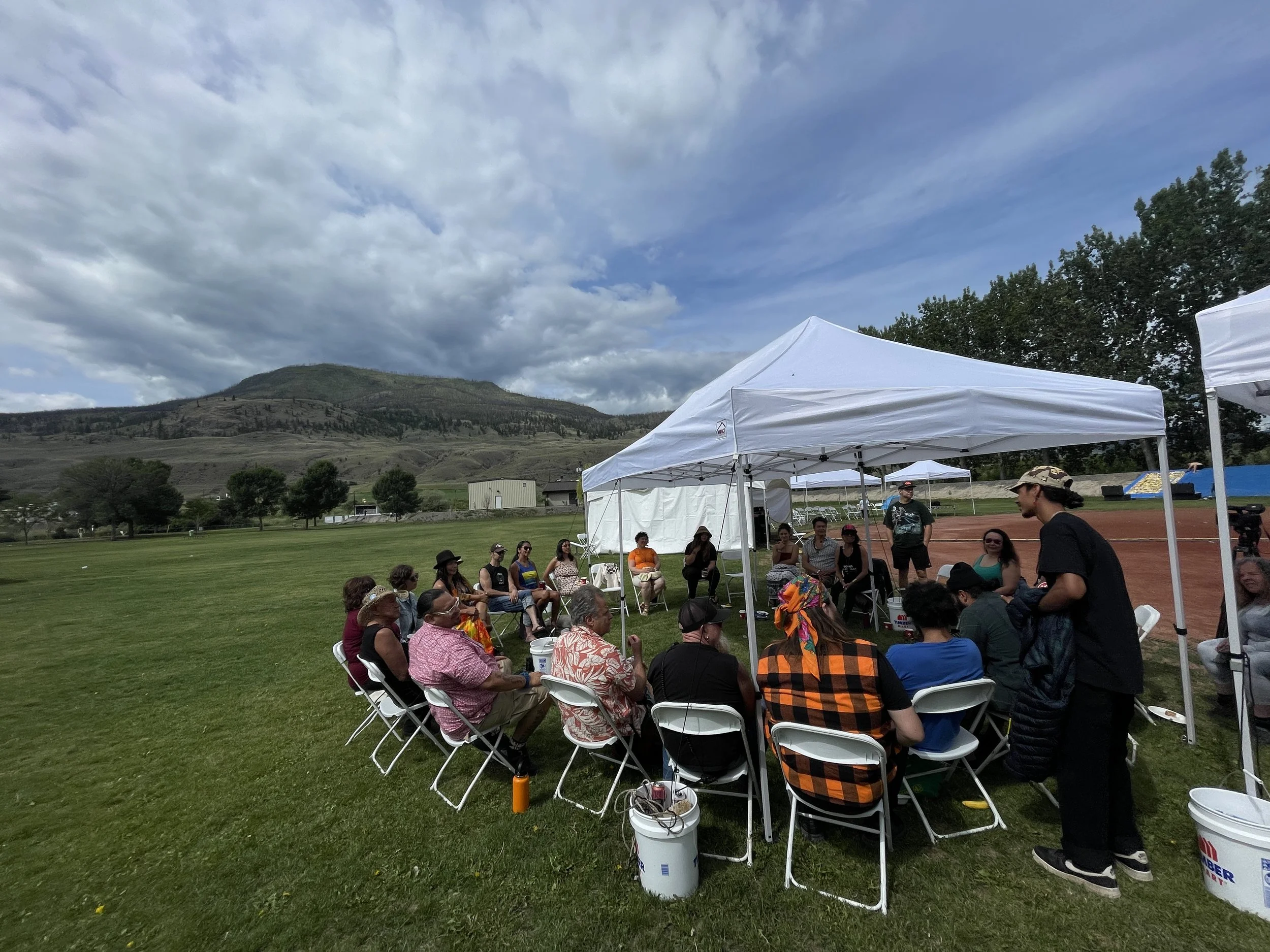 An outdoor photo of a group of people sitting in a circle, on chairs in a grassy field. There is a large hill in the background. The sky looks dramatic, with a mix of thick and whispy clouds on a blue sky.