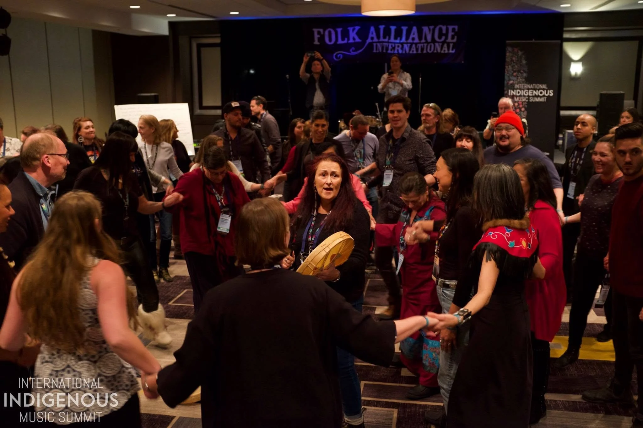 Sandy Scofield plays a hand drum while people dance hand-in-hand around her in a banquet room at a hotel in New Orleans.