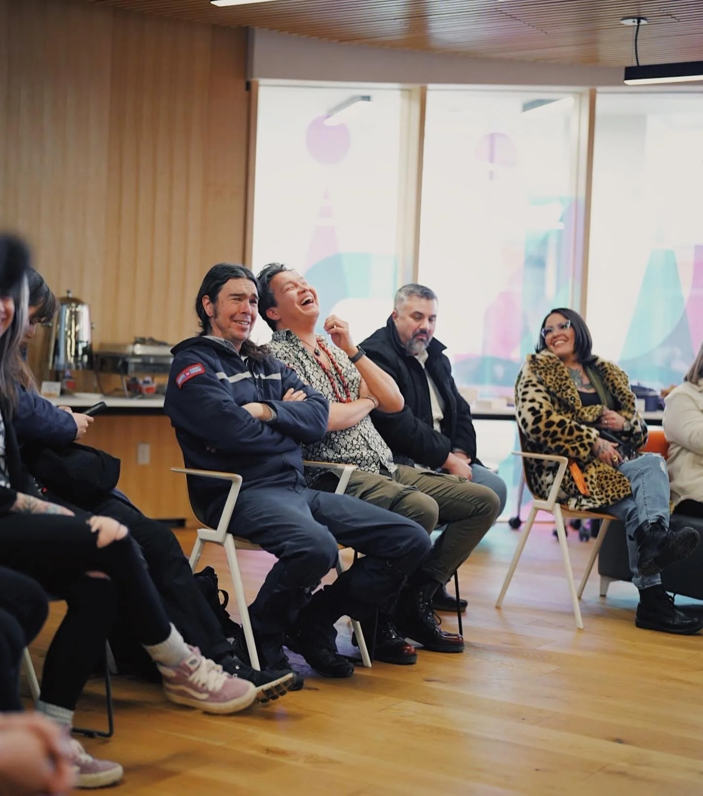 A candid photo of a few people sitting in chairs at a gathering in a hotel meeting room. The main 2 male subjects can be seen laughing.