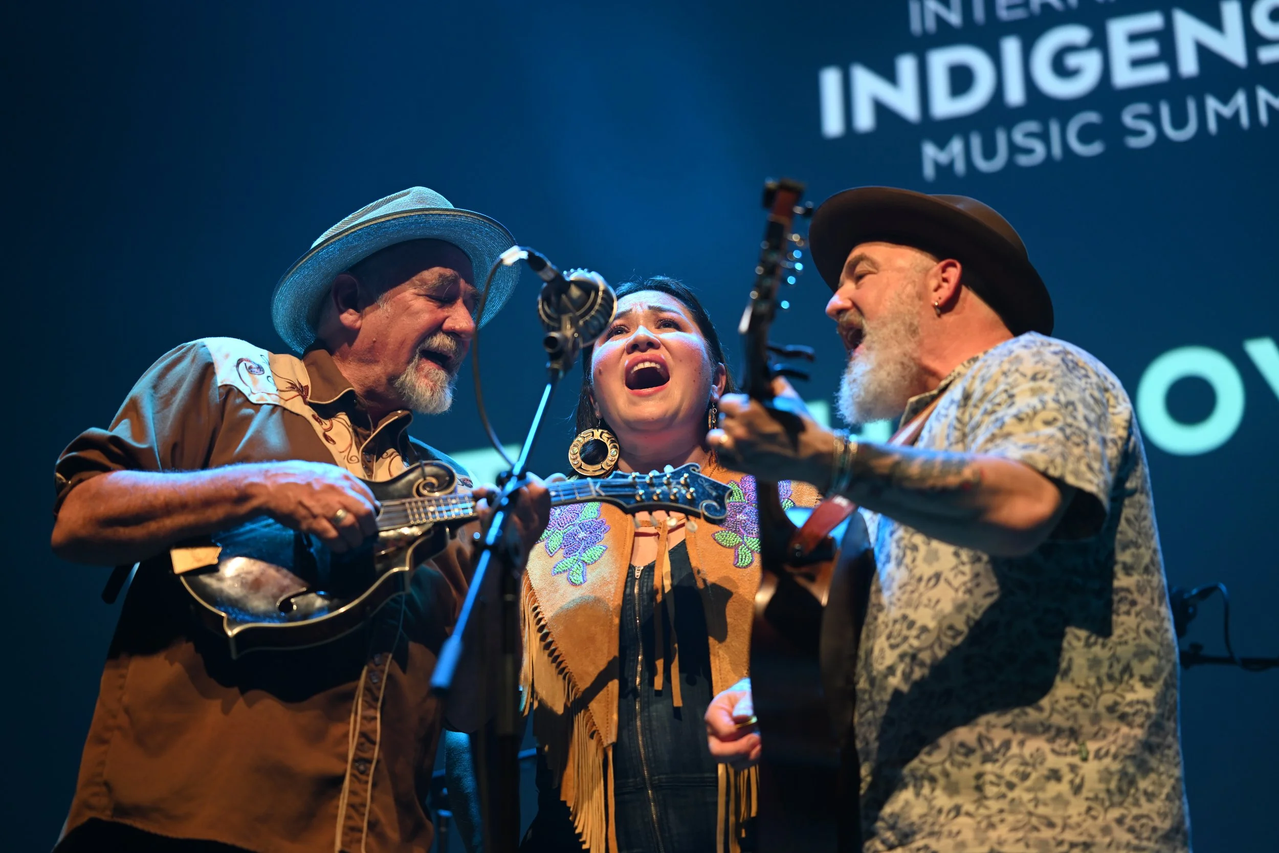 A performance photo of Diyet & The Love Soldiers singing into the microphone. One is playing mandolin, one singing, one playing guitar.