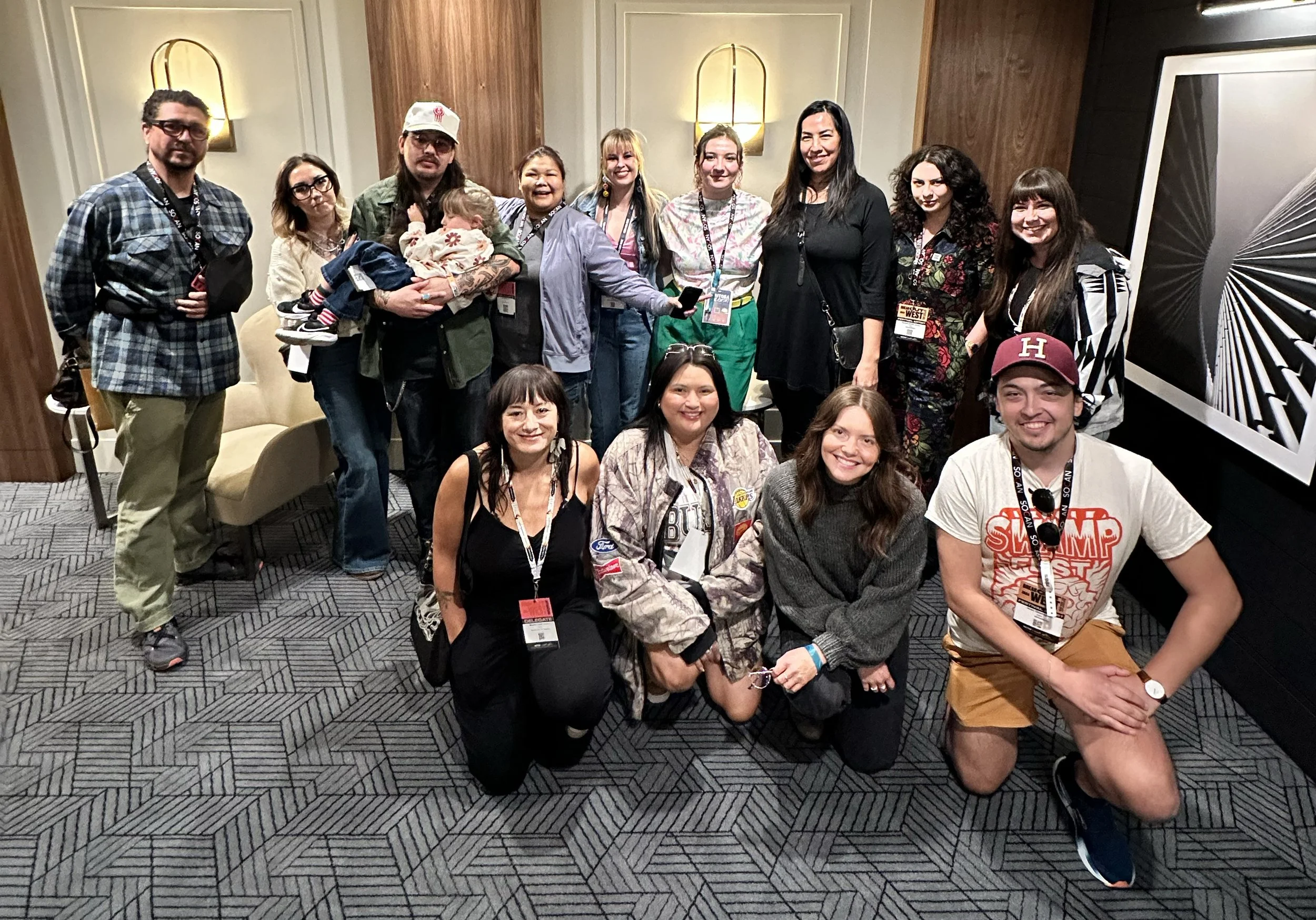 A group photo of 10-15 people standing, some kneeling, in a hotel hallway, smiling at the camera.