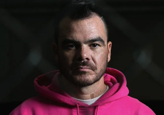 A photo of Graeme Jones looking serious at the camera, wearing a pink hoodie.
