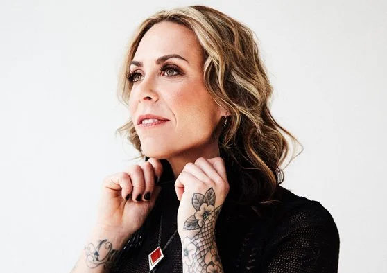 A photo of Amanda Rheaume holding her collar, looking away from the camera. She has blonde wavy hair and a floral tattoo on her lower left arm.